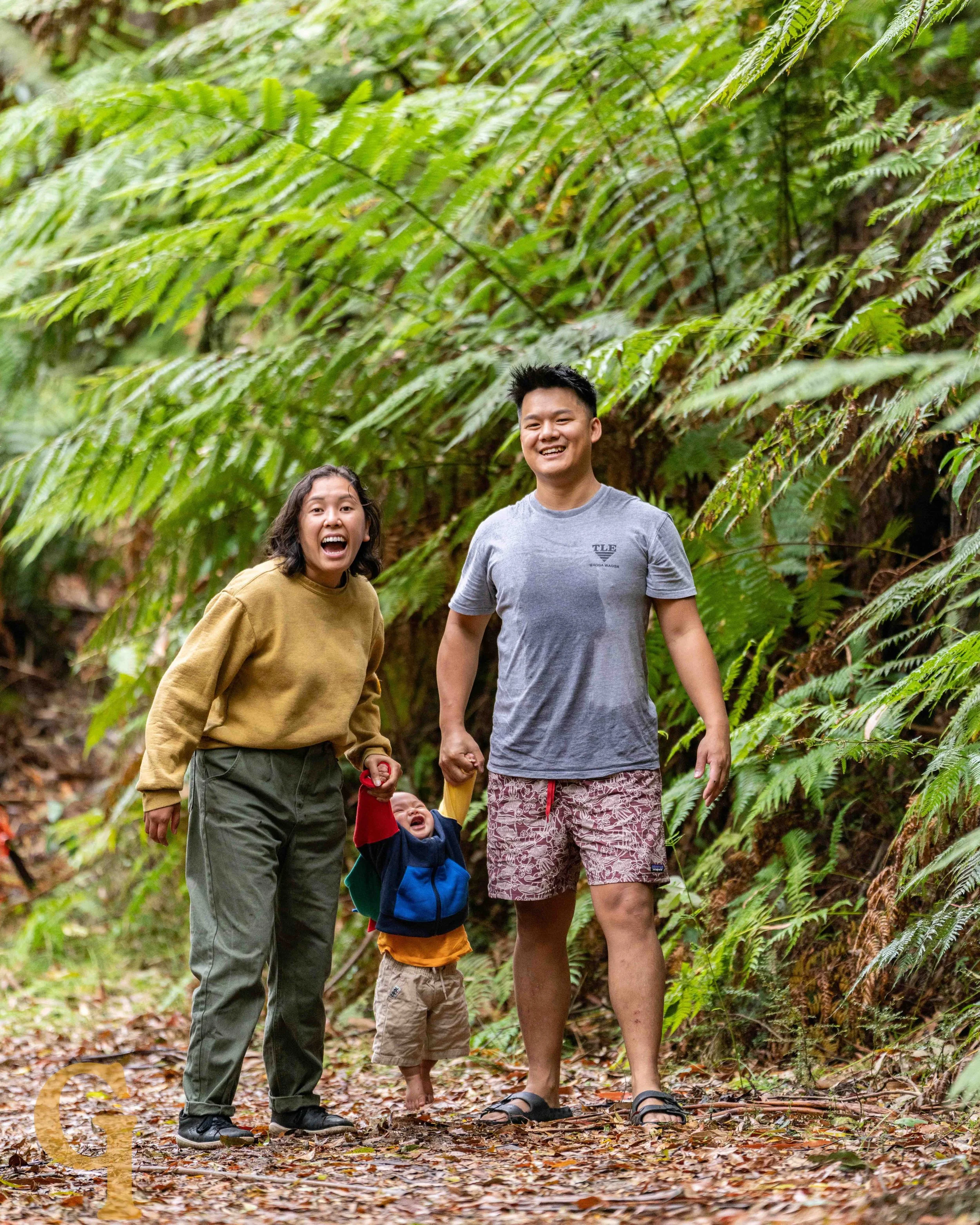 A family of three, including a woman, a man, and a toddler, enjoying a walk through a lush green rainforest. The woman and man are smiling and the toddler is being held by the woman. Ian Grant is skilled in a variety of portrait photography.