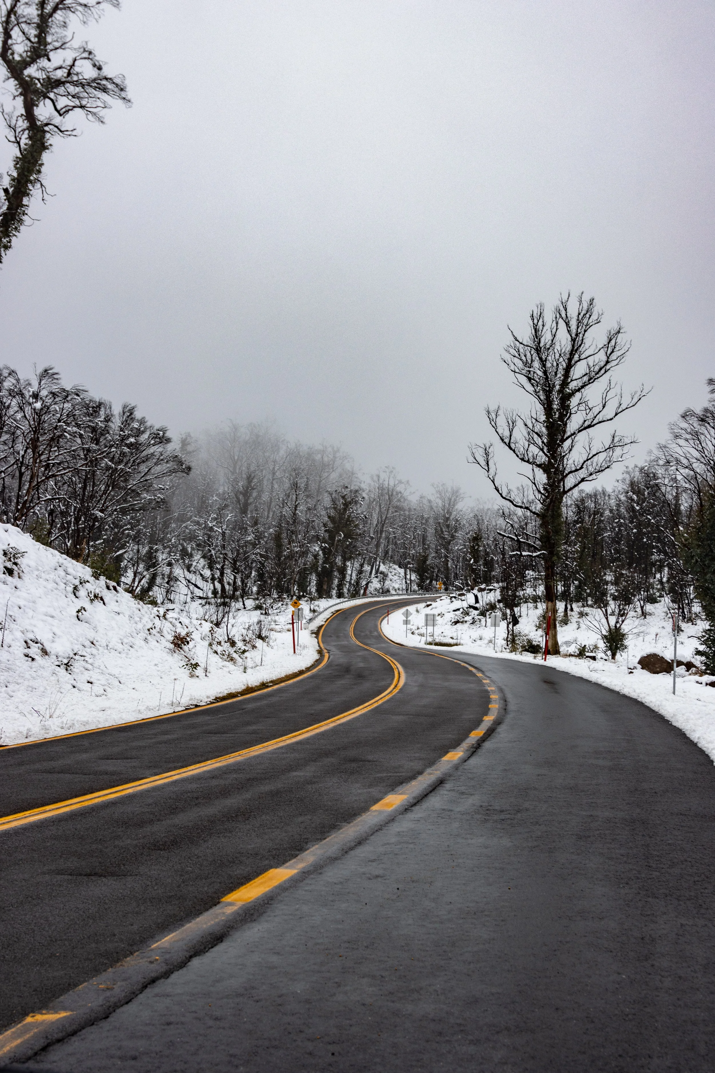 Winding mountain road with snow on sides and leafless trees, overcast sky.