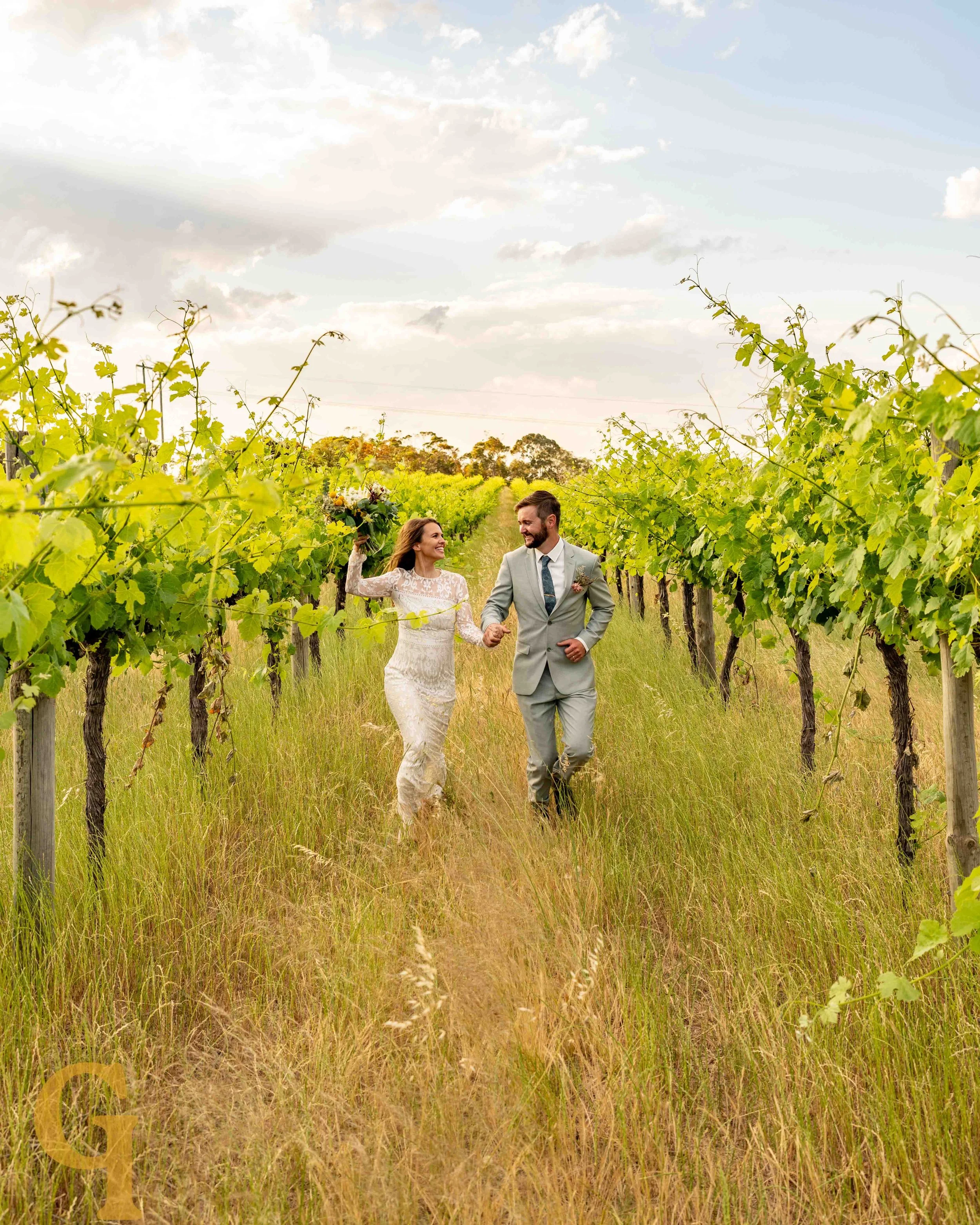 A bride and groom walking hand-in-hand through a vineyard on their wedding day, with the bride holding a bouquet and smiling at each other on a sunny day.