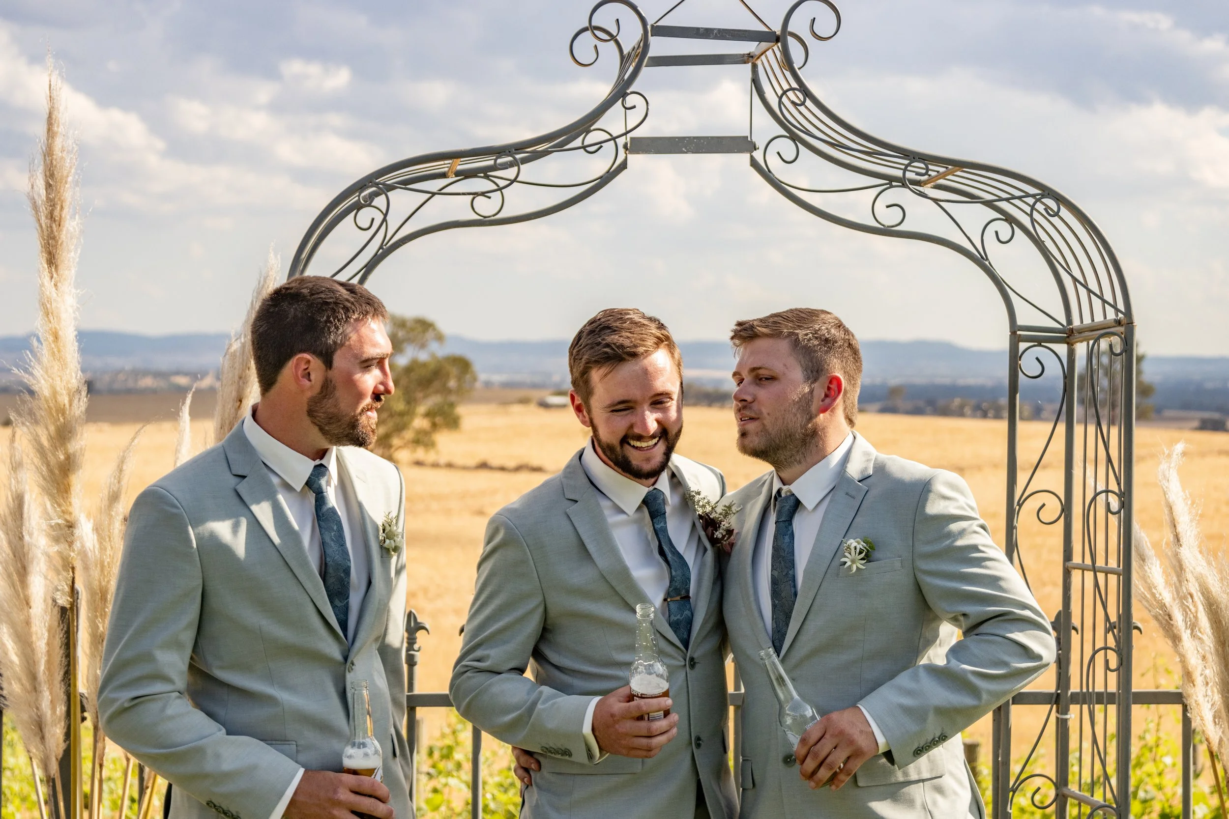 Three men in light gray suits and dark ties, standing under a metal archway, holding beer bottles, at an outdoor wedding in a countryside field with mountains in the background.