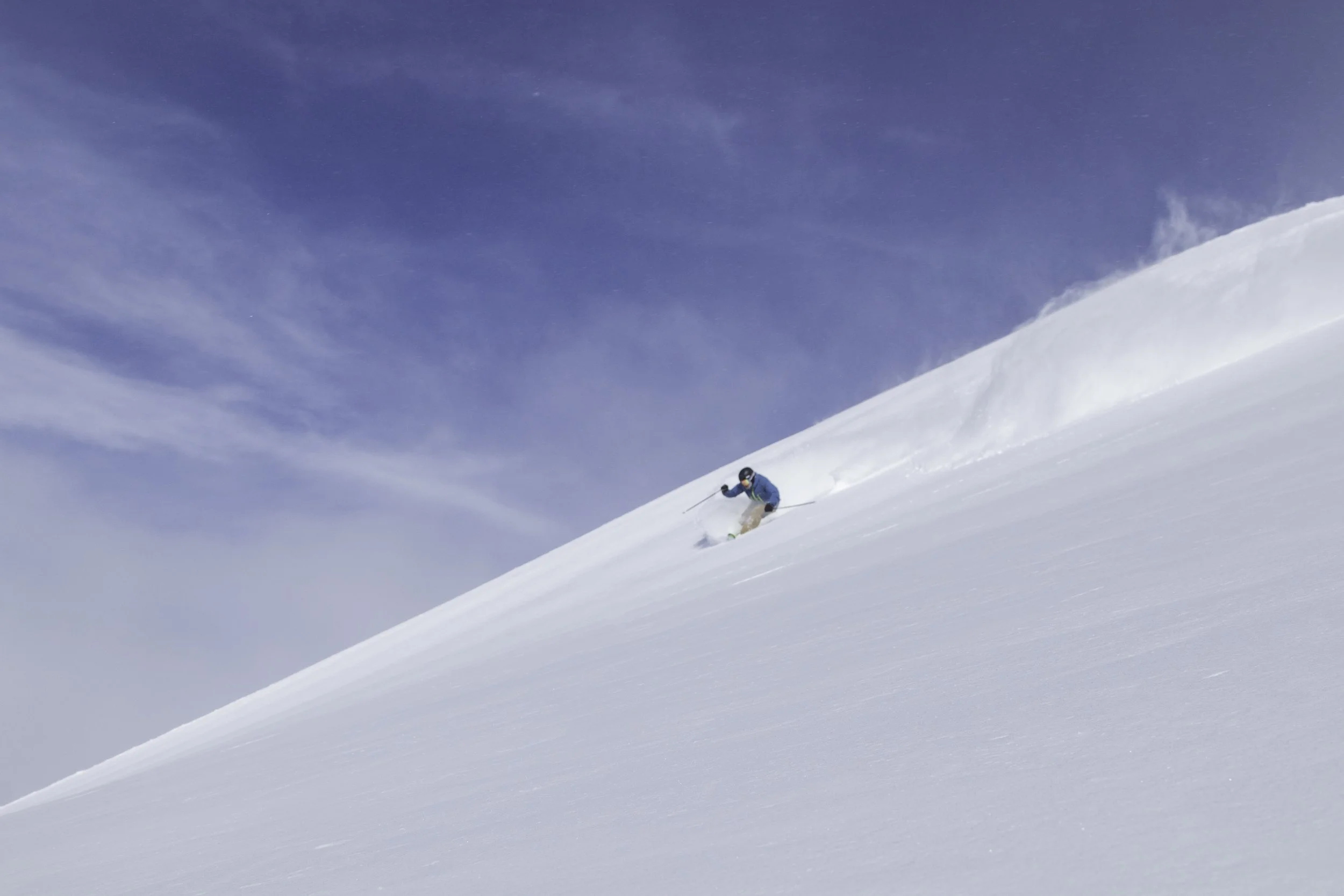 A skier in blue and black gear skiing down a snow-covered slope on a clear, sunny day.