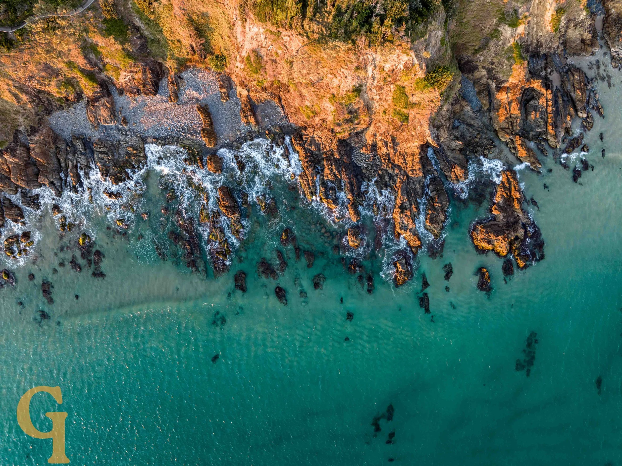 An aerial view of a rugged coastline with rocky cliffs, green vegetation, and turquoise ocean waters with waves crashing against the rocks.