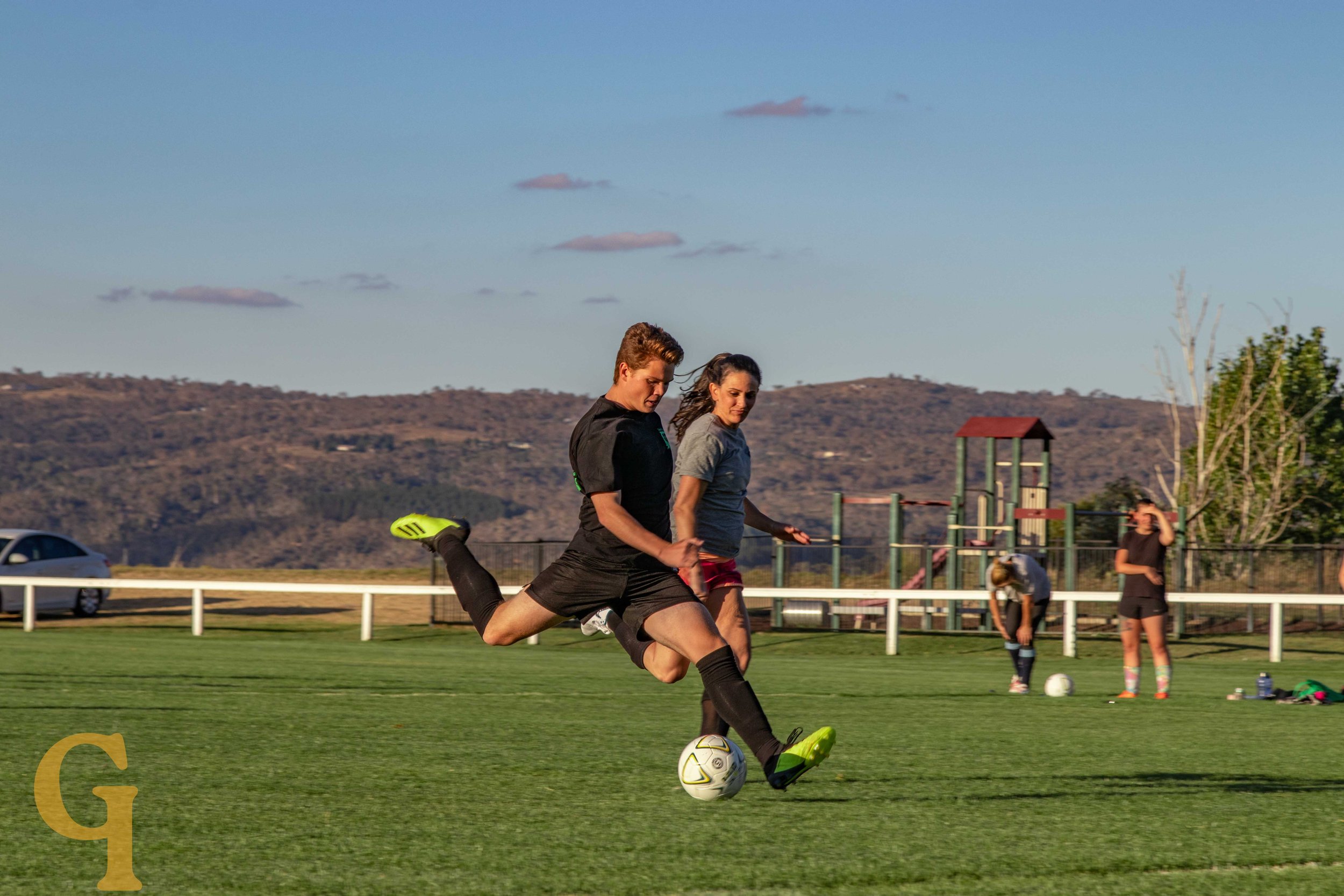 A male and female soccer player in black and gray shirts compete for the ball on a grass field, with a playground, trees, and mountains in the background.