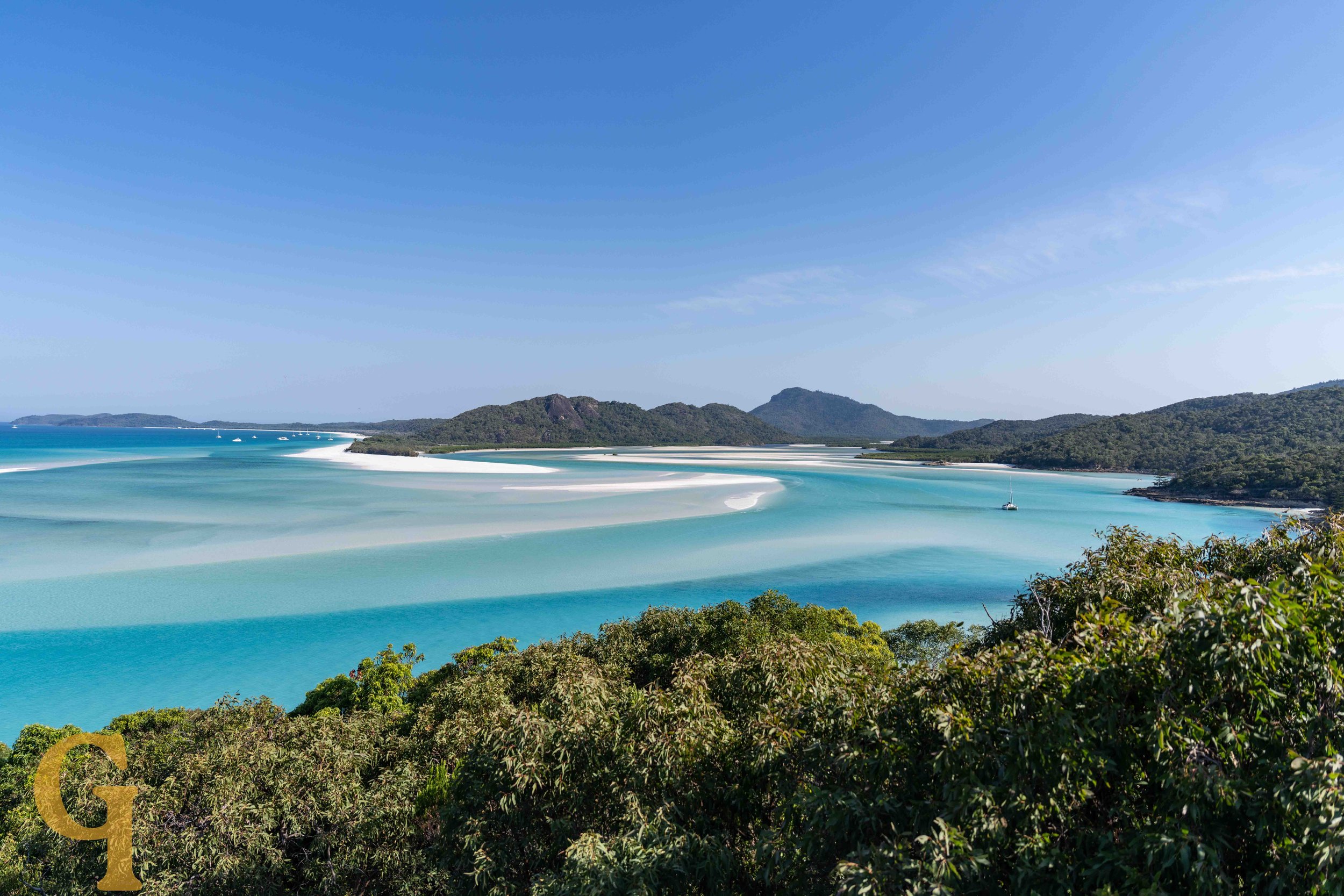 A scenic view of a beach with white sandbars, turquoise water, green islands in the background, and clear blue sky.