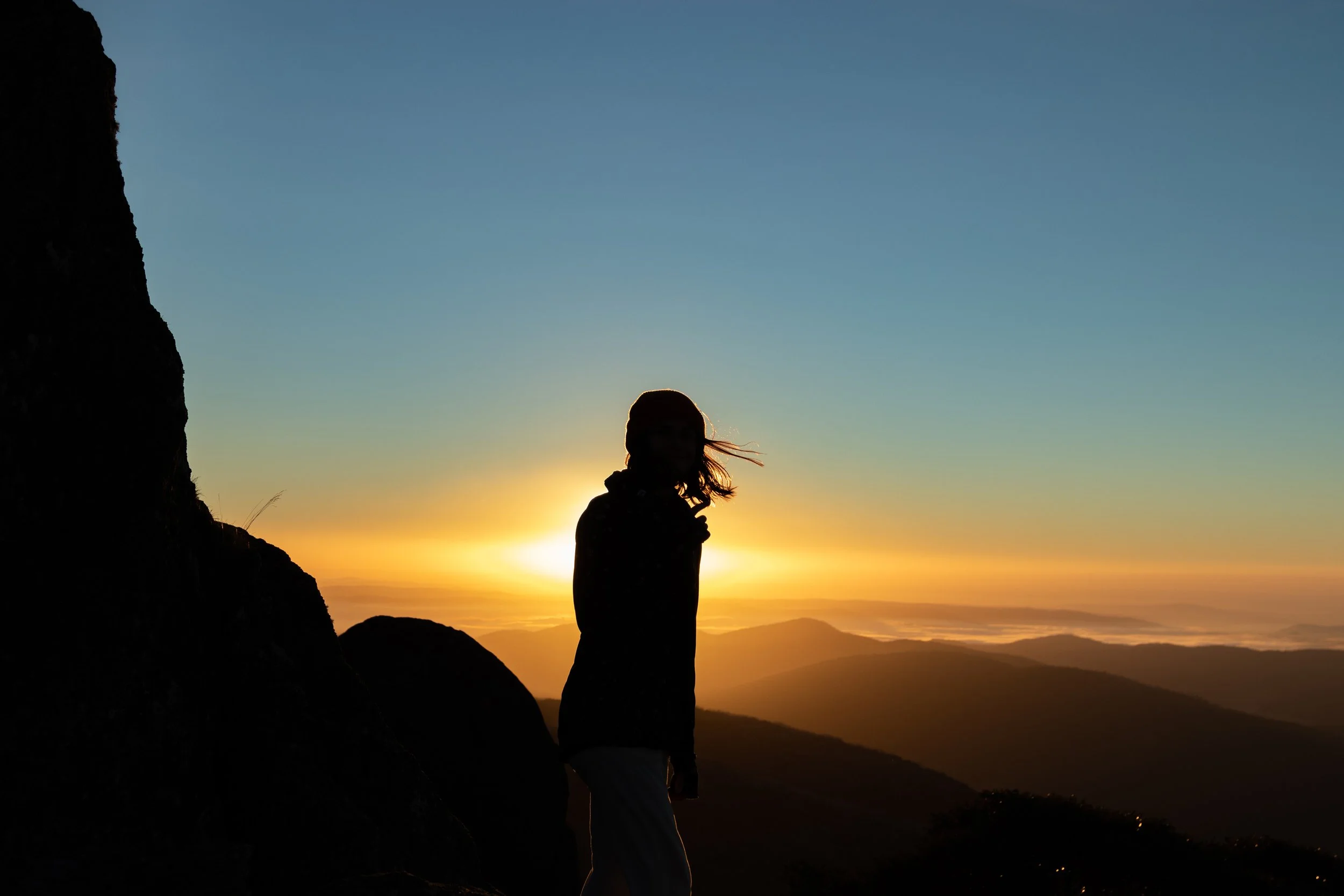 A person standing on a mountain at sunset with a backdrop of mountains and a colorful sky.