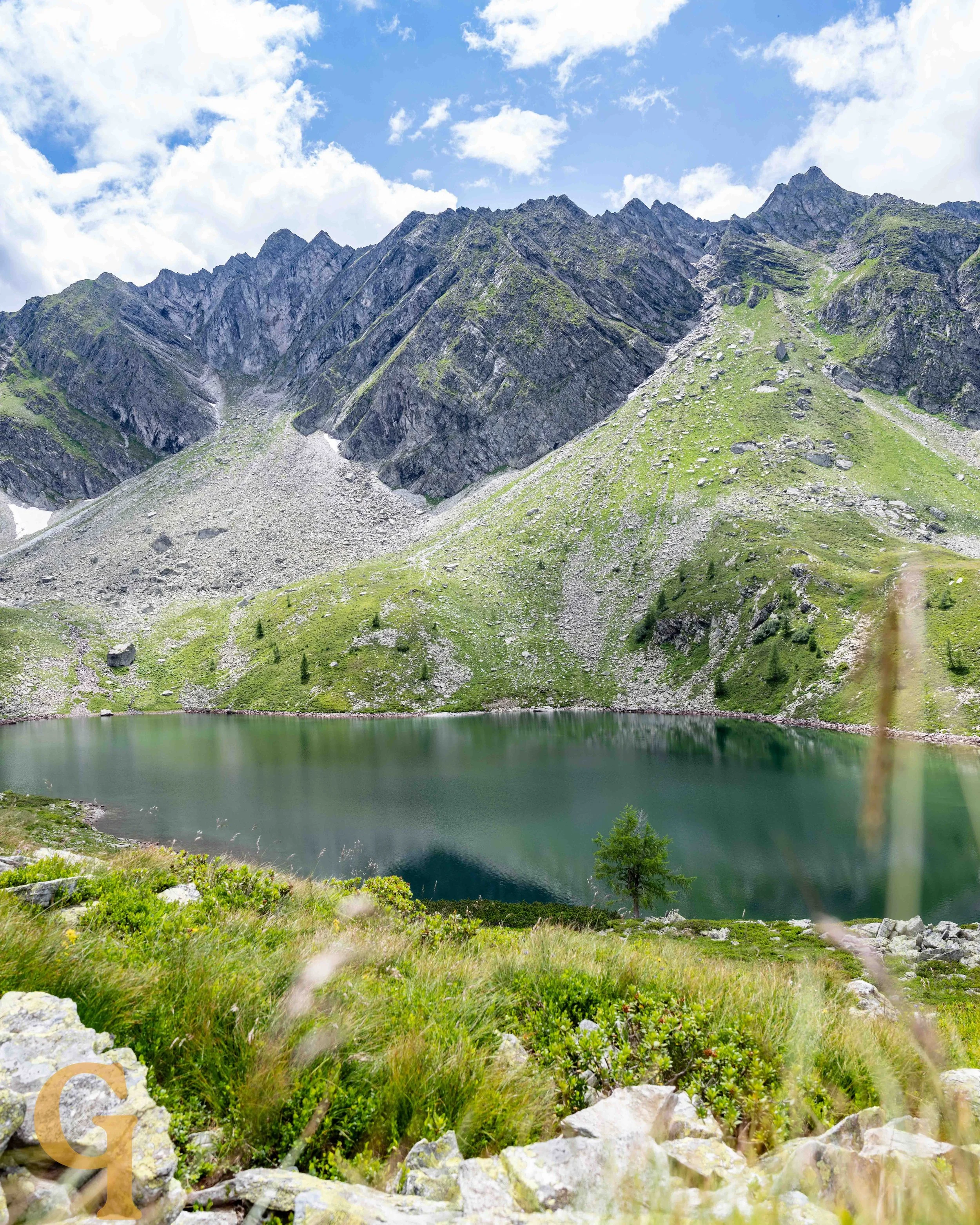 A mountain landscape with rocky peaks, green slopes, and a small lake at the base, under a partly cloudy sky.