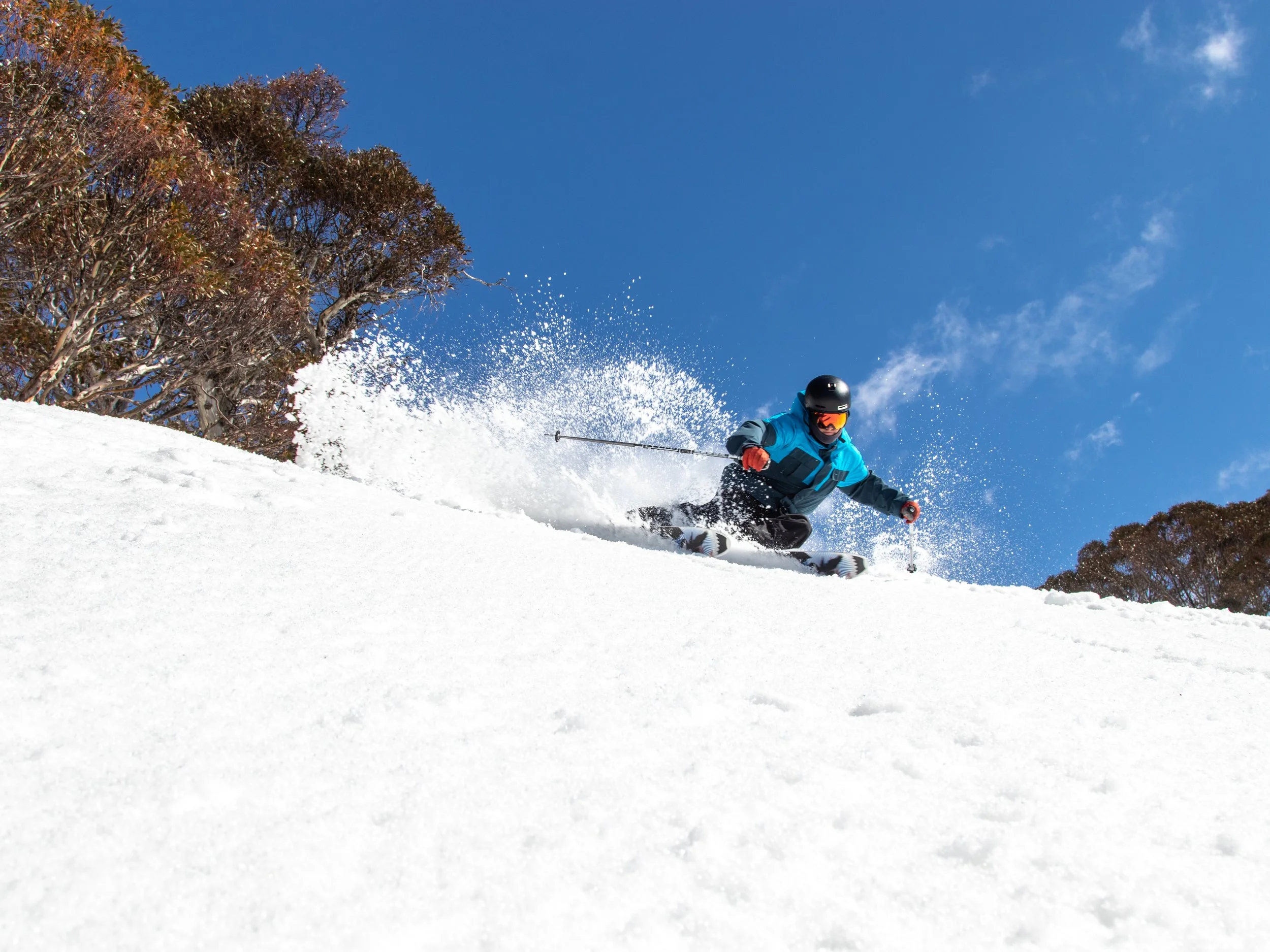 A skier wearing a blue jacket, black helmet, and orange goggles skiing down a snowy slope under a clear blue sky with trees in the background.