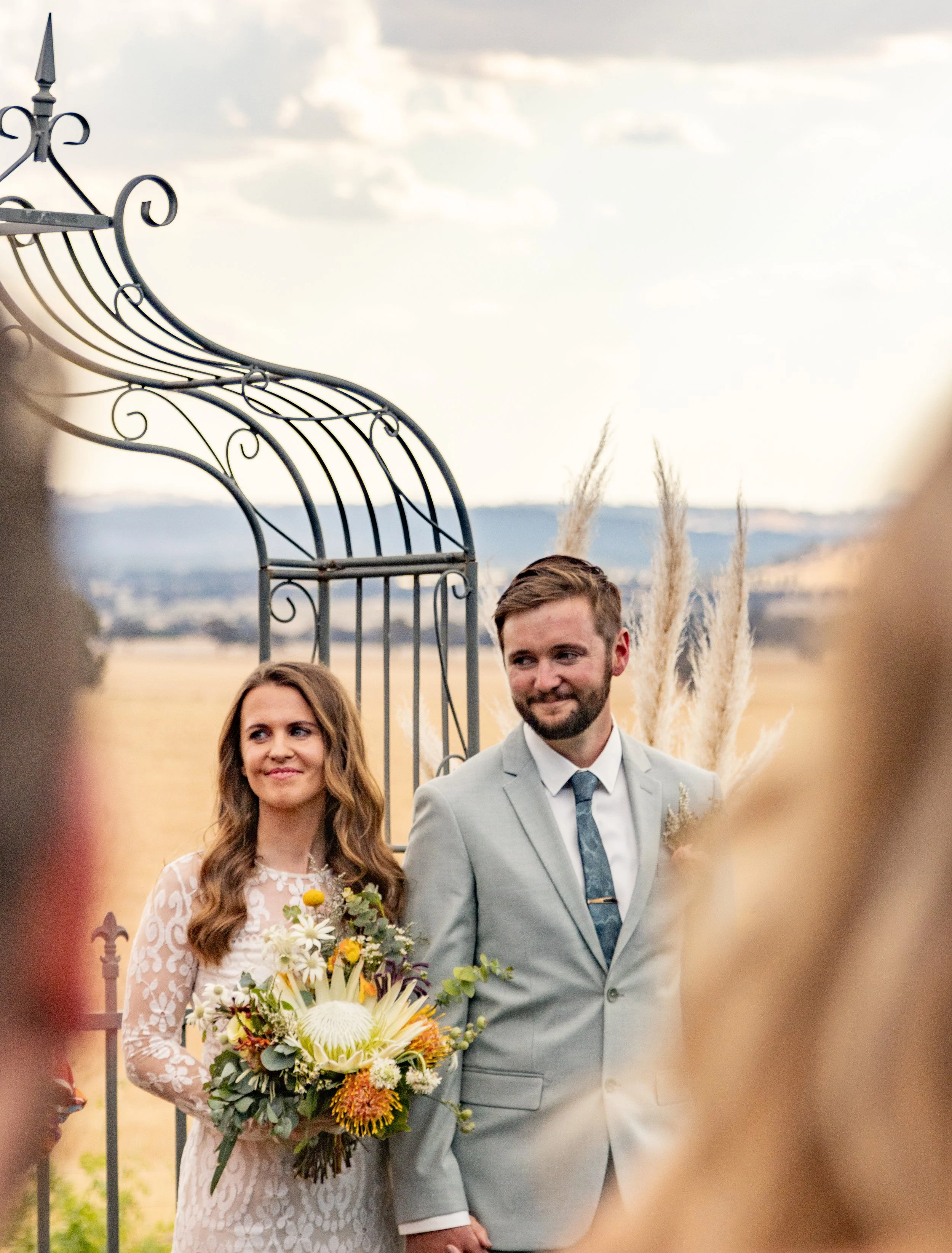 Bride holding a bouquet standing next to groom in a gray suit during outdoor wedding ceremony.