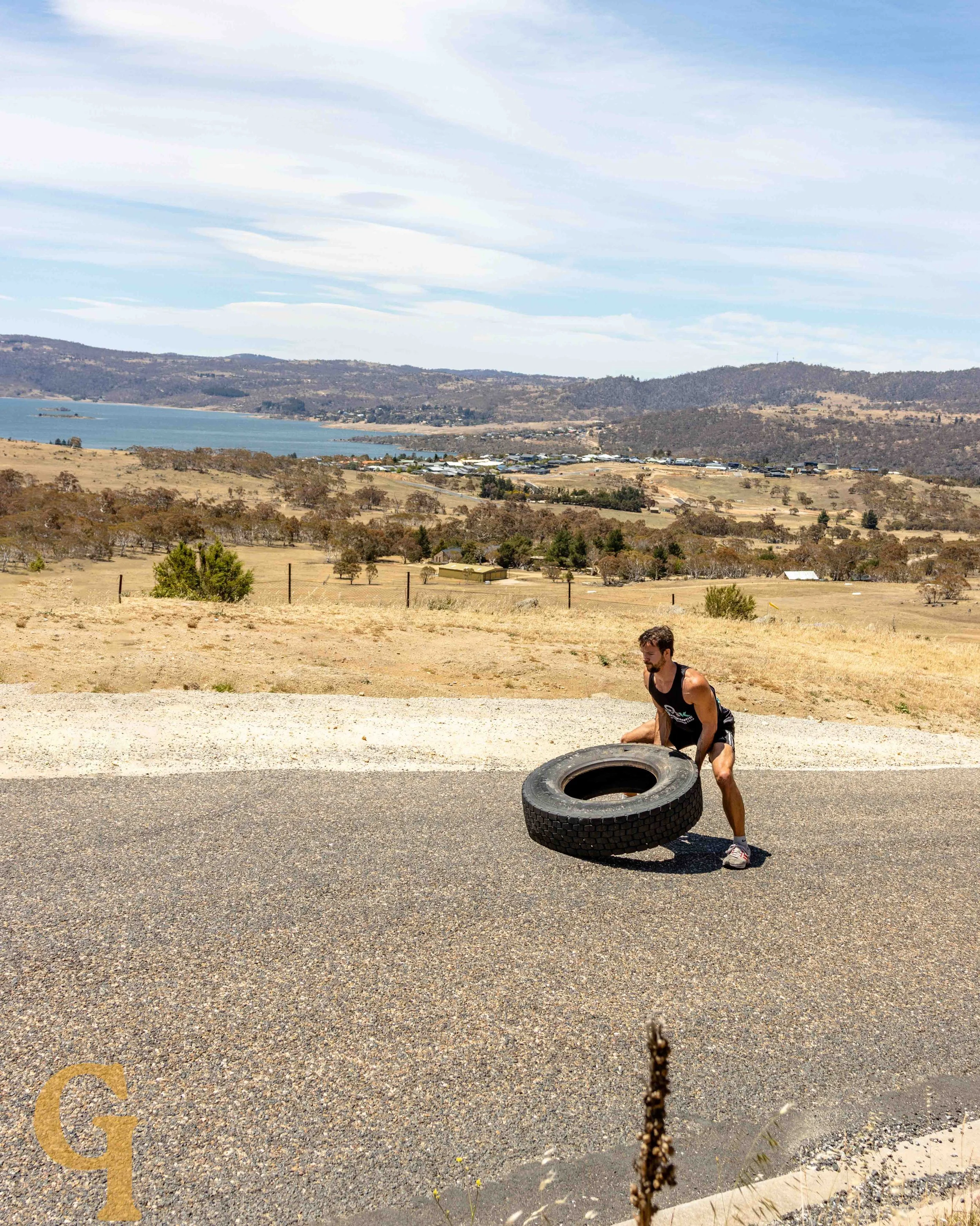 Man lifting a large tire outdoors on a gravel surface with rolling hills, a lake, and mountains in the background under a partly cloudy sky.