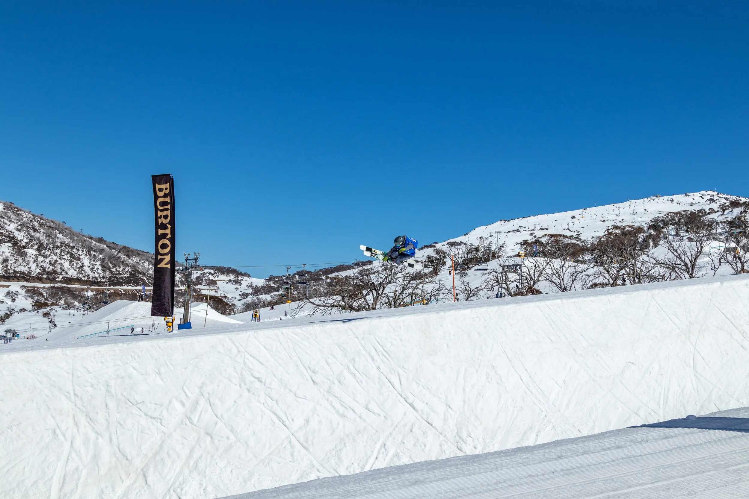 A skier in blue gear performing a jump over a snow ramp at a ski resort with snow-covered mountains and clear blue sky in the background.