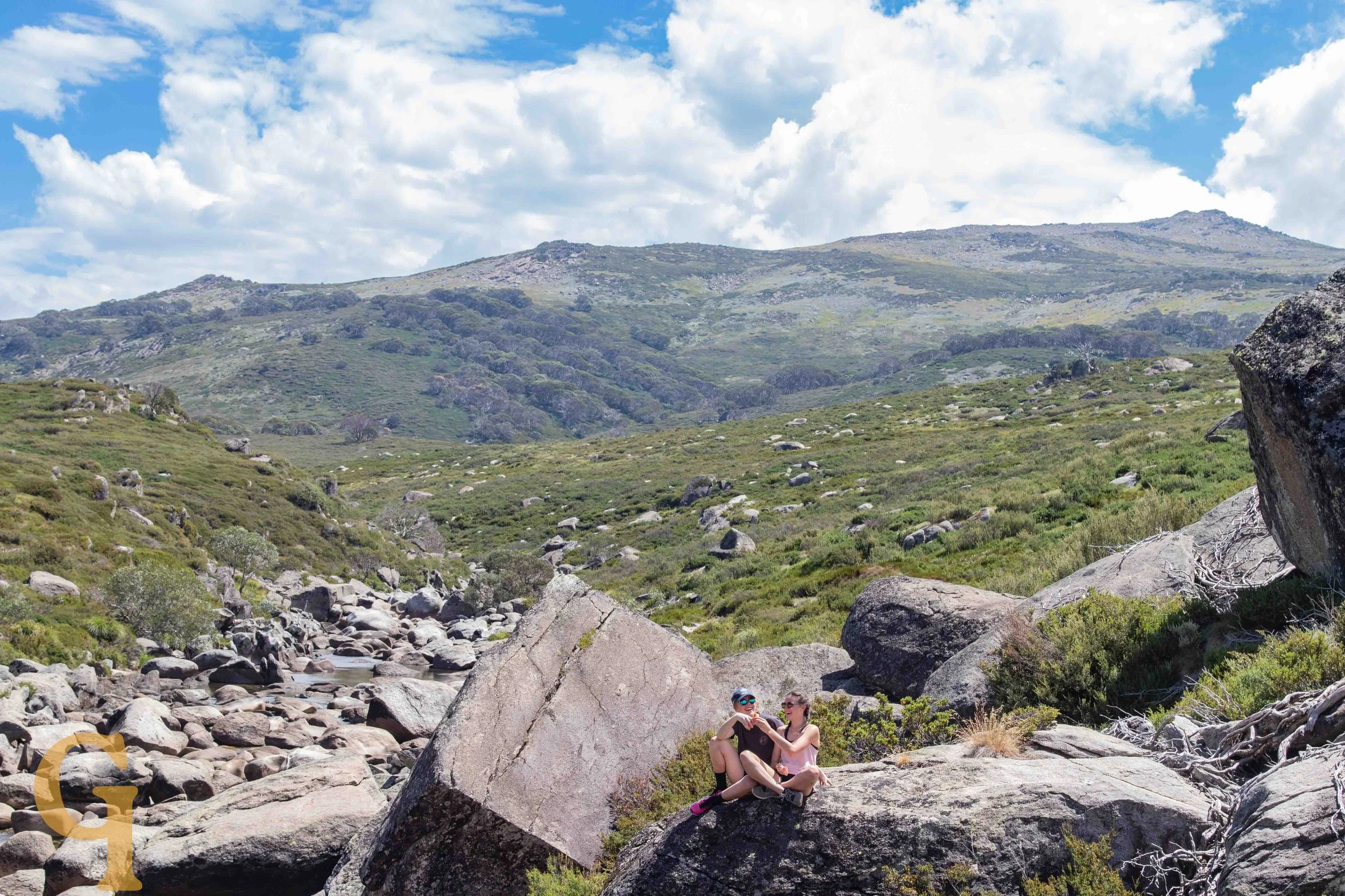 Two people sitting on a large rock in a mountainous landscape with green hills, rocks, and a partly cloudy sky.