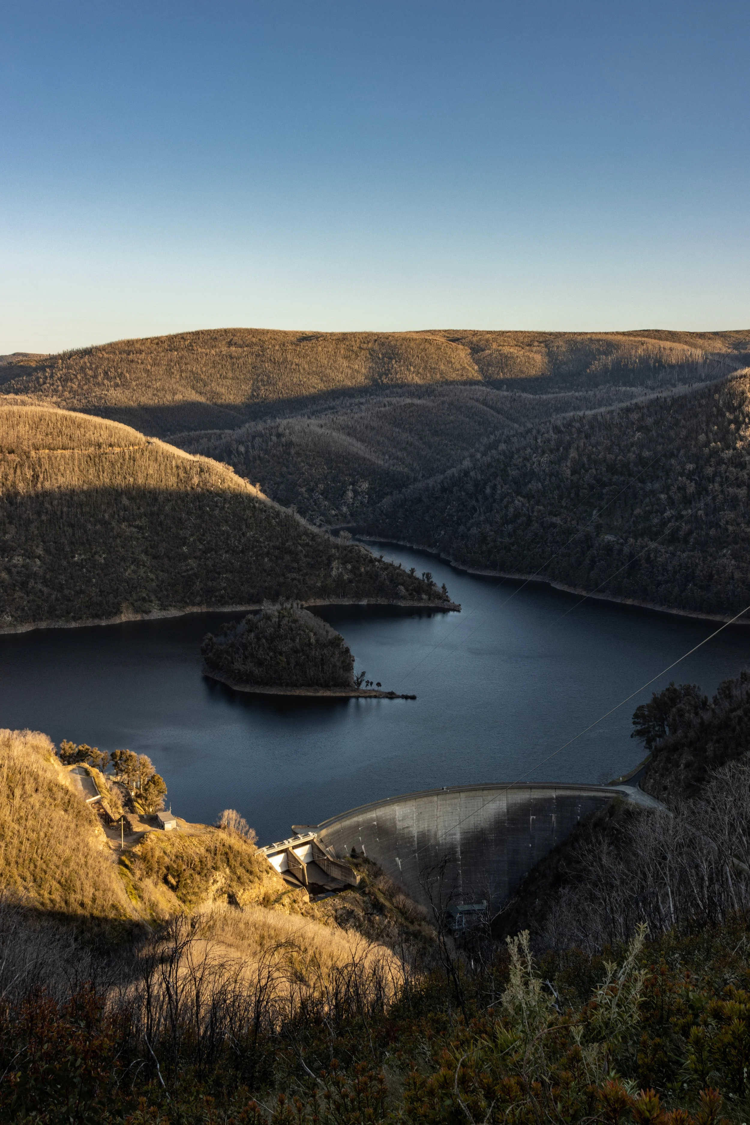 A dam with water surrounded by hills and mountains under a clear blue sky.