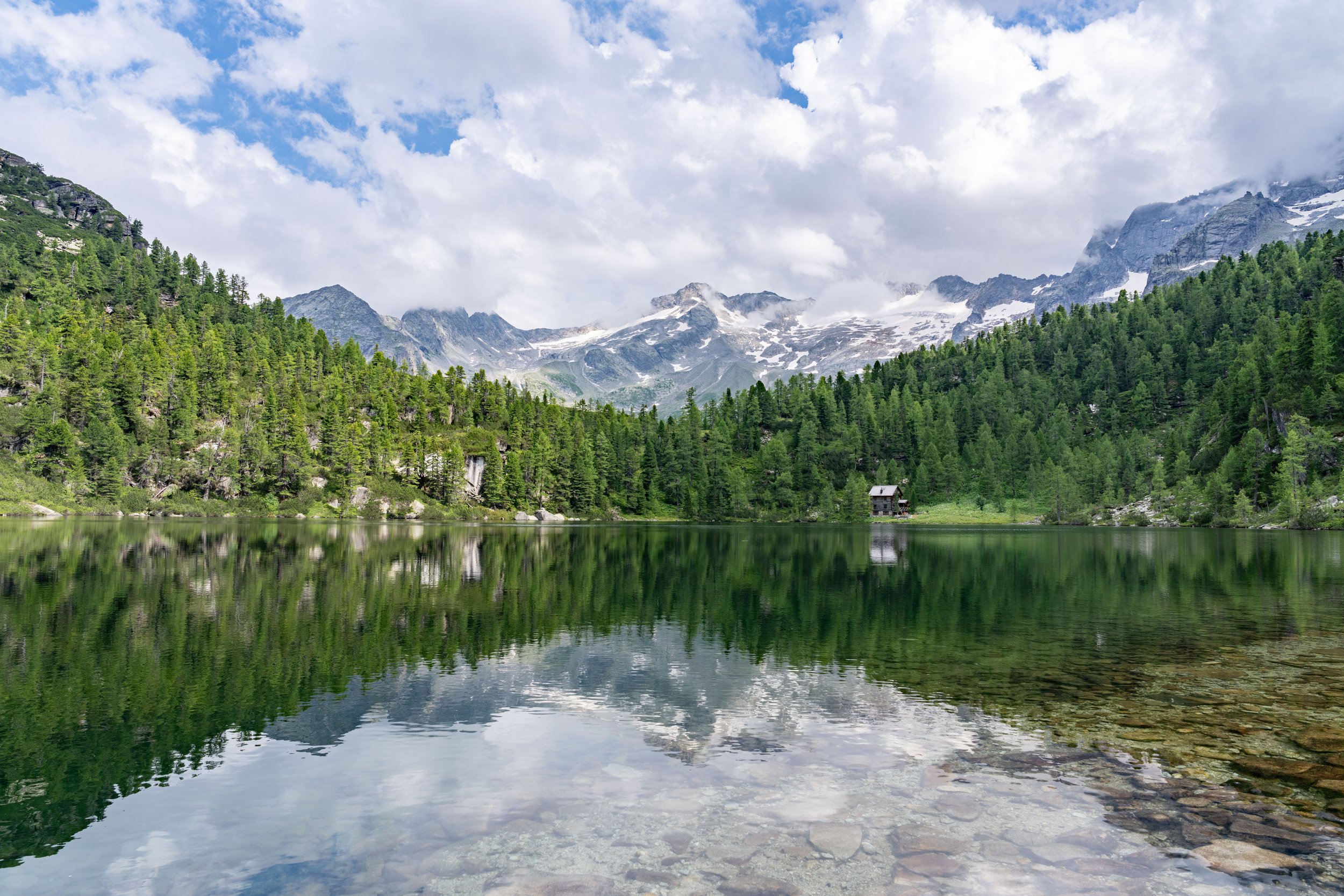 Scenic view of a mountain lake surrounded by dense green trees with snow-capped mountains in the background under partly cloudy sky.