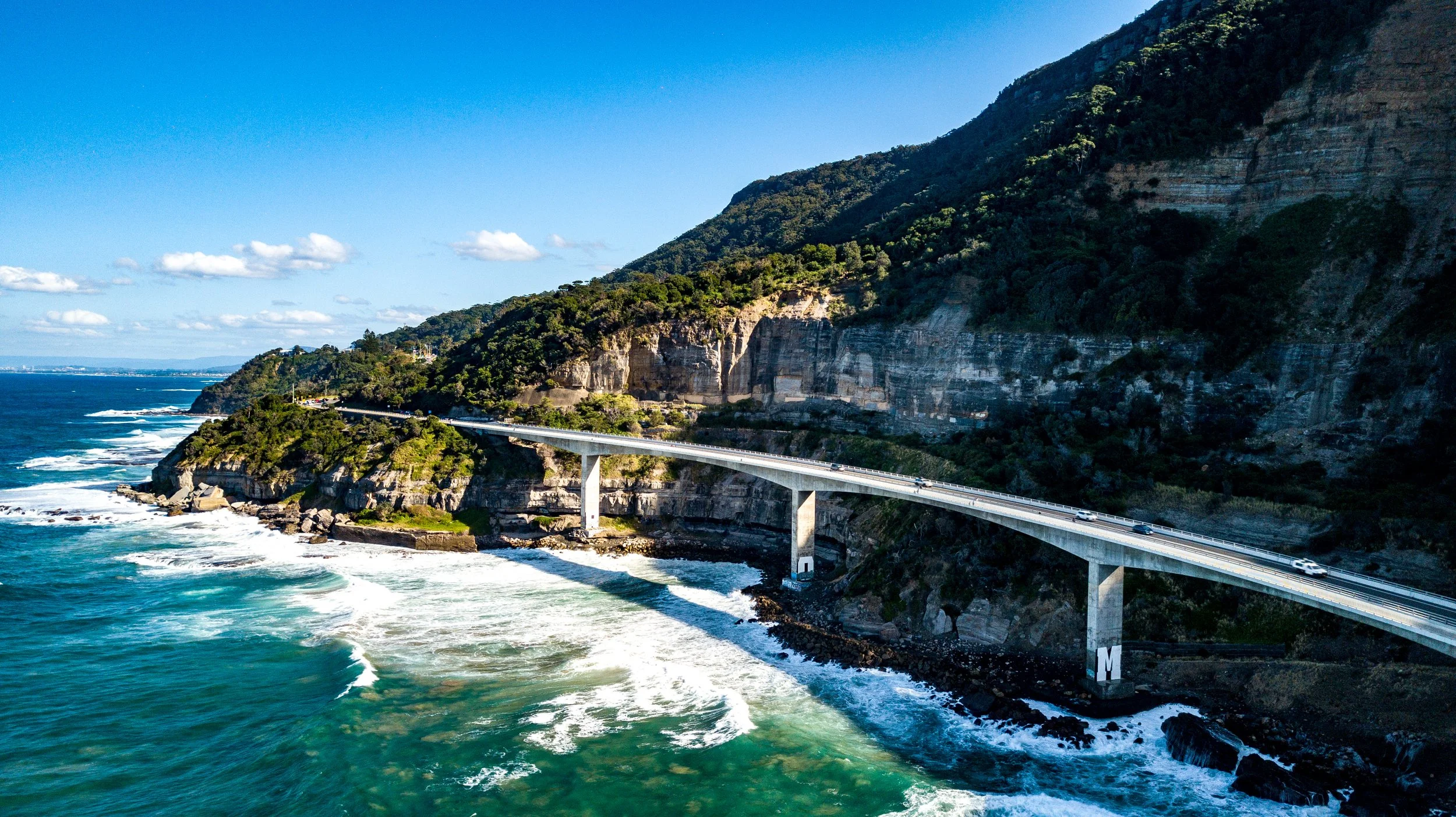A coastal highway bridge stretched over the ocean with waves crashing below, alongside rocky cliffs and a forested hillside under a clear blue sky.