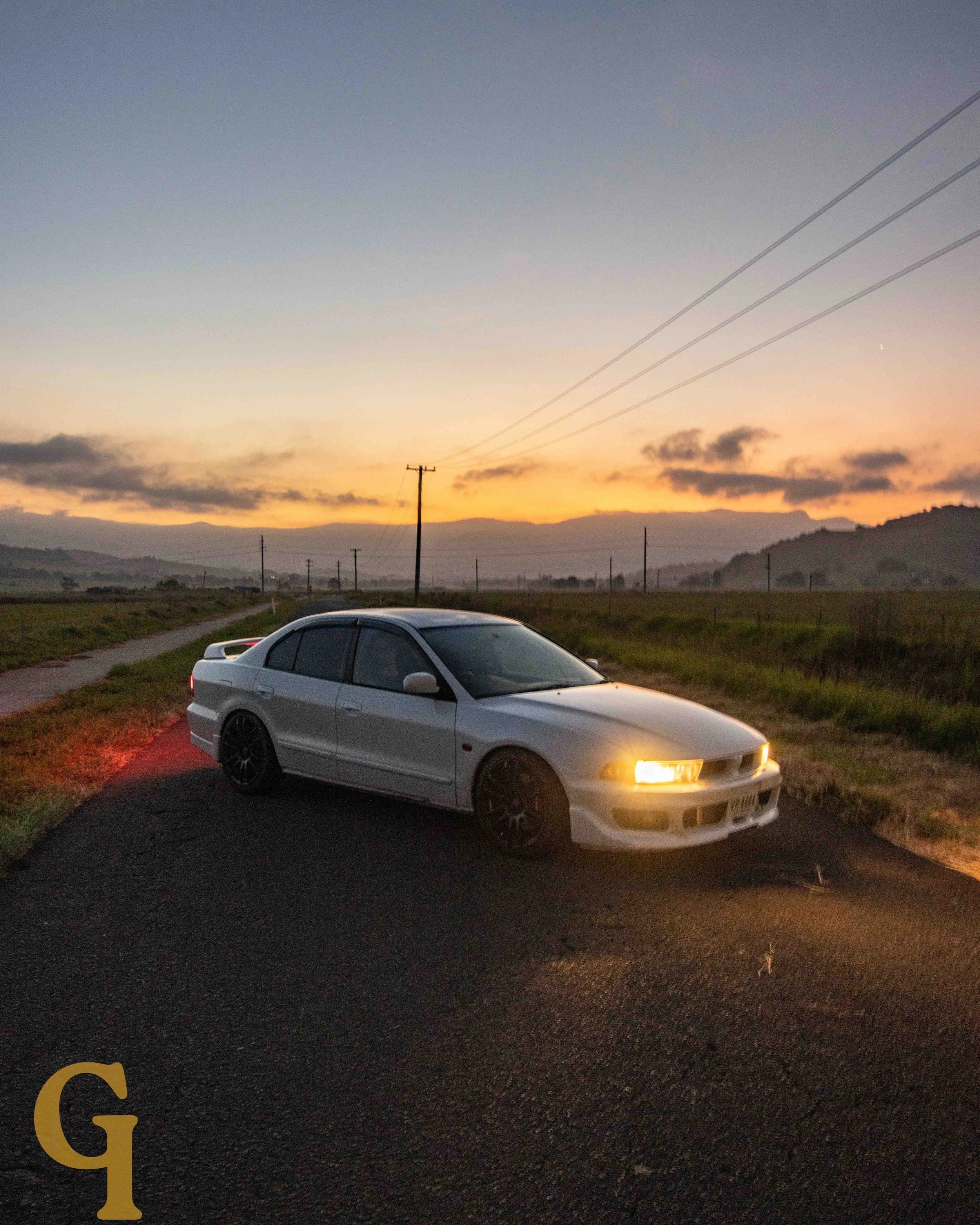 White sedan car with illuminated headlights parked on side of gravel road at sunset in rural landscape.