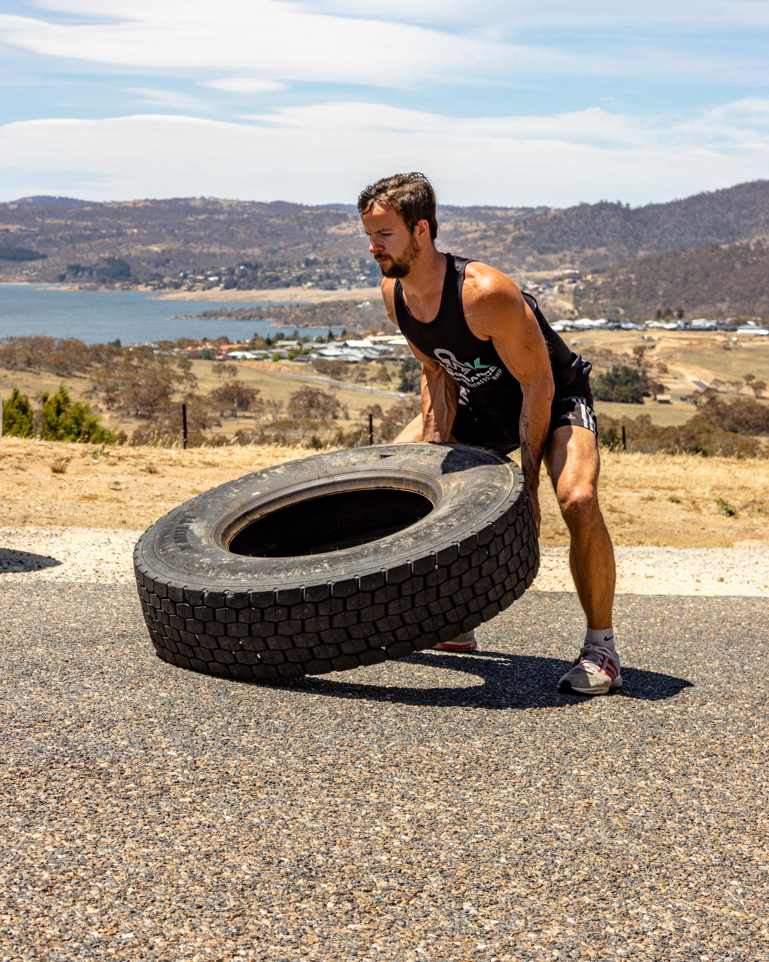 A man in athletic attire lifting a large tire outdoors on a sunny day with hills, a lake, and a rural landscape in the background.