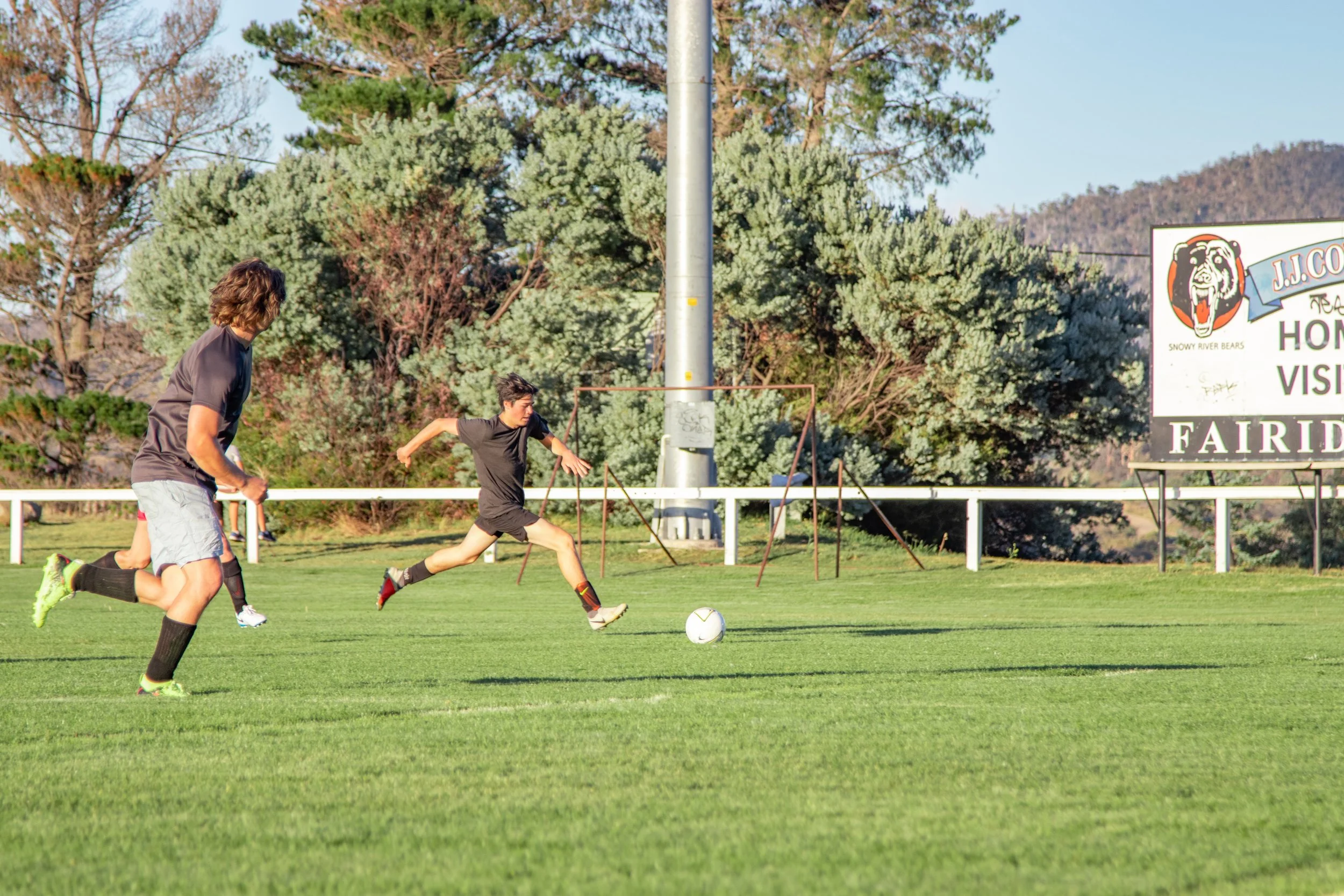 Two young men playing soccer on a grassy field with a goalpost in the background, surrounded by trees.