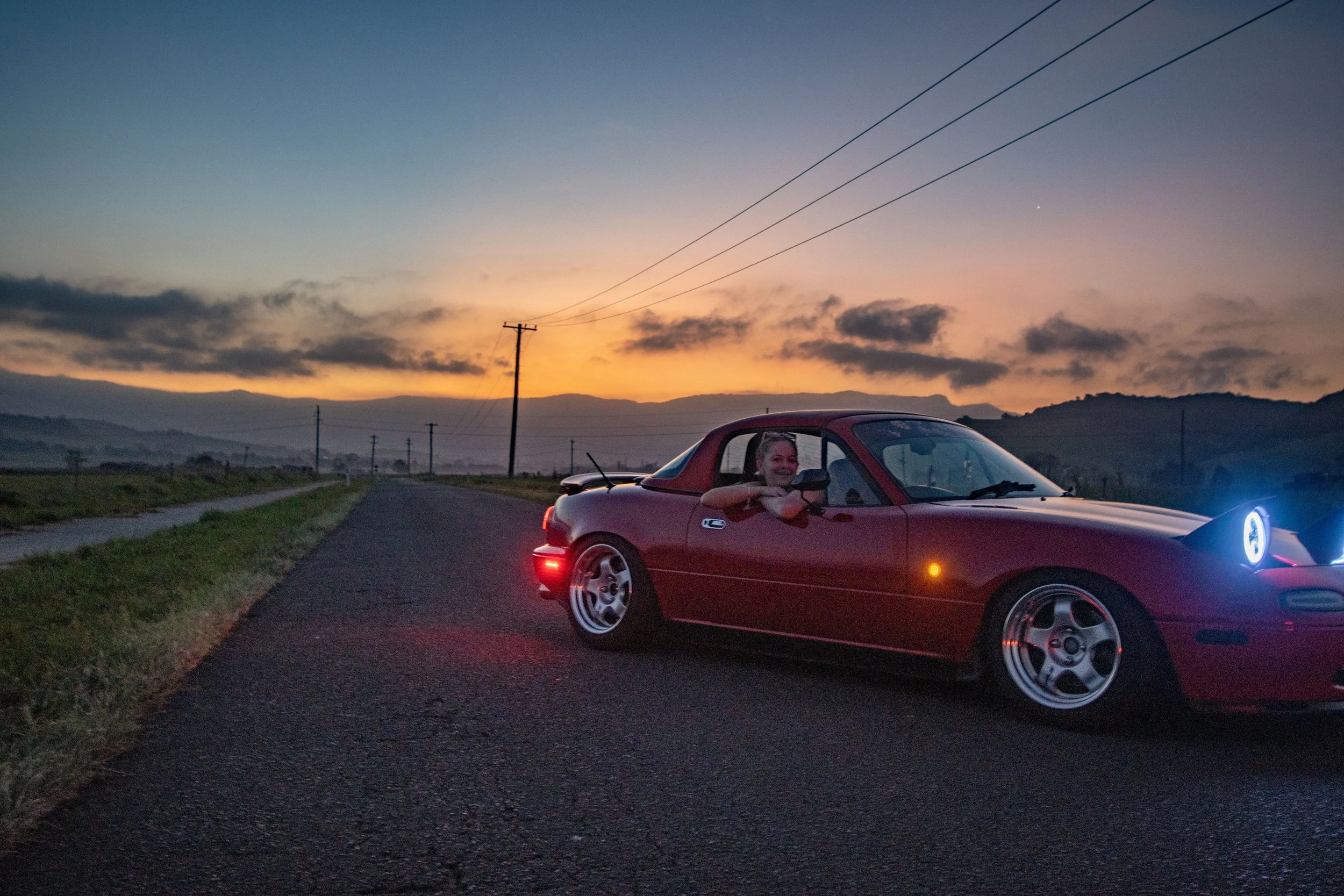 A woman smiling and leaning out of the driver's side window of a red sports car on a rural road at sunset with hills and power lines in the background.