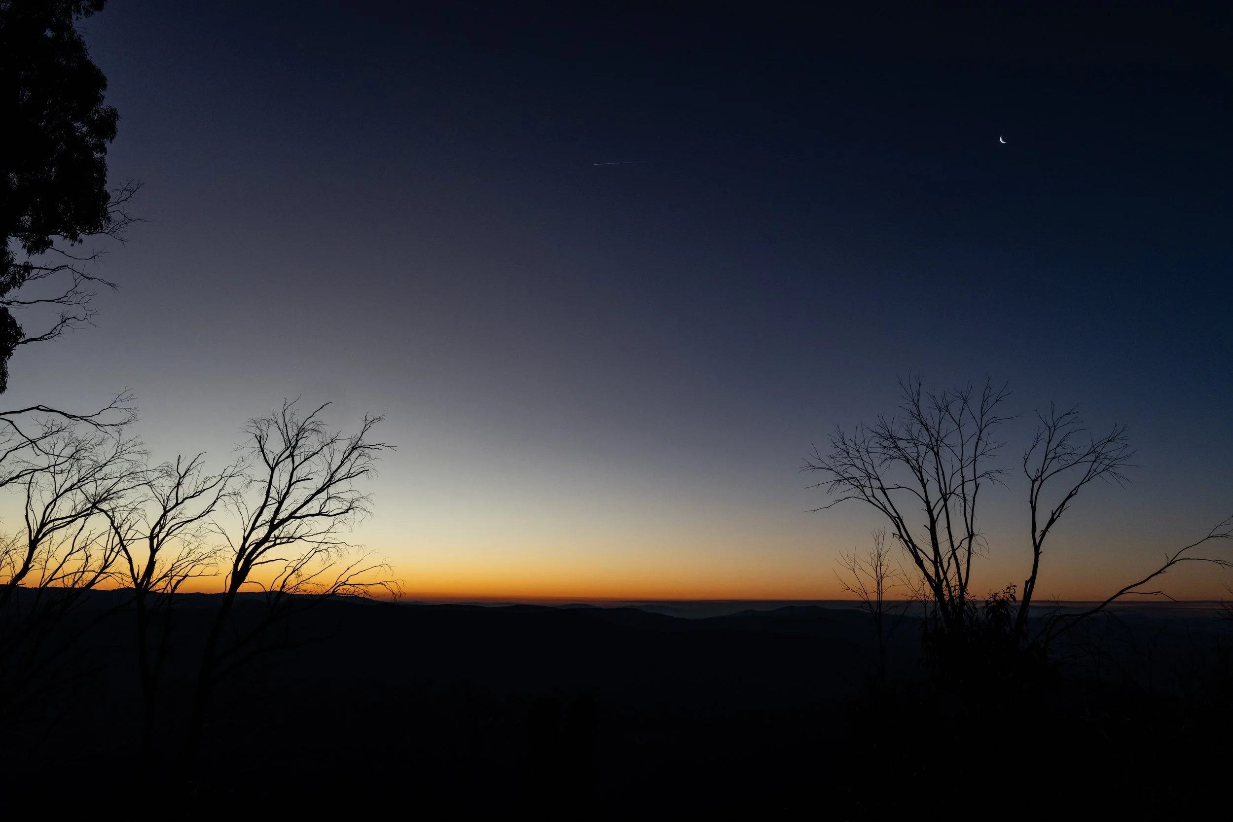 A landscape scene at dusk with a dark sky, a narrow crescent moon, a faint star, and a streak of light. Silhouettes of leafless trees are in the foreground, and distant mountains are visible on the horizon.