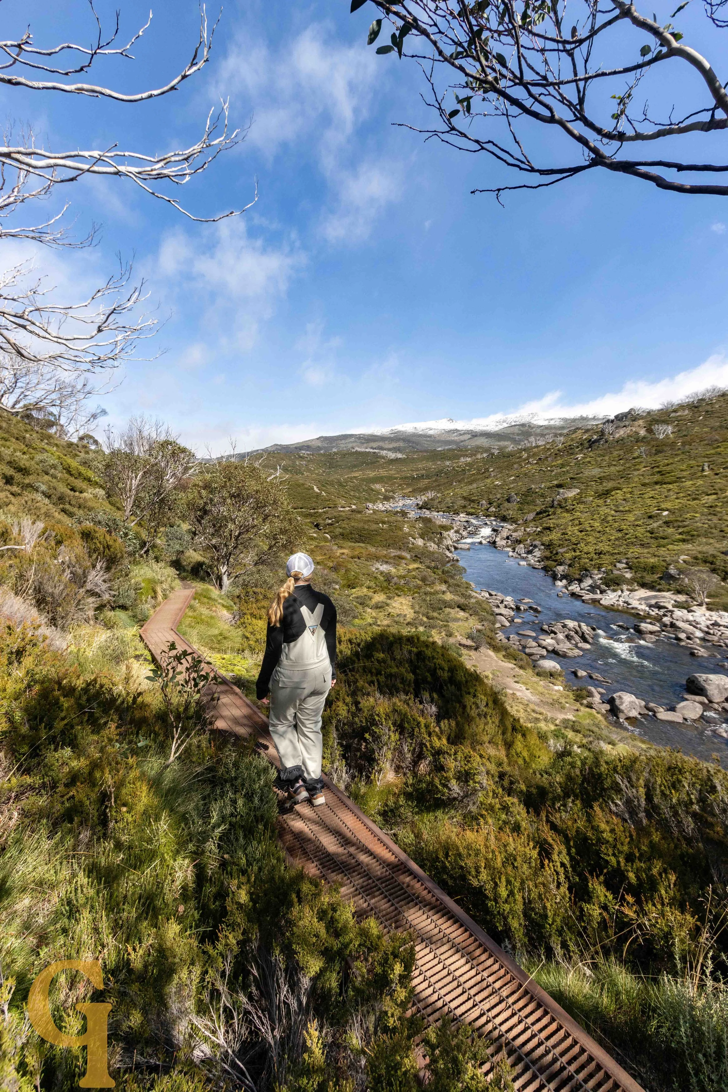 A person walking on a narrow, rusty metal railway track through green shrubland near a flowing river with rocks, under a partly cloudy blue sky.