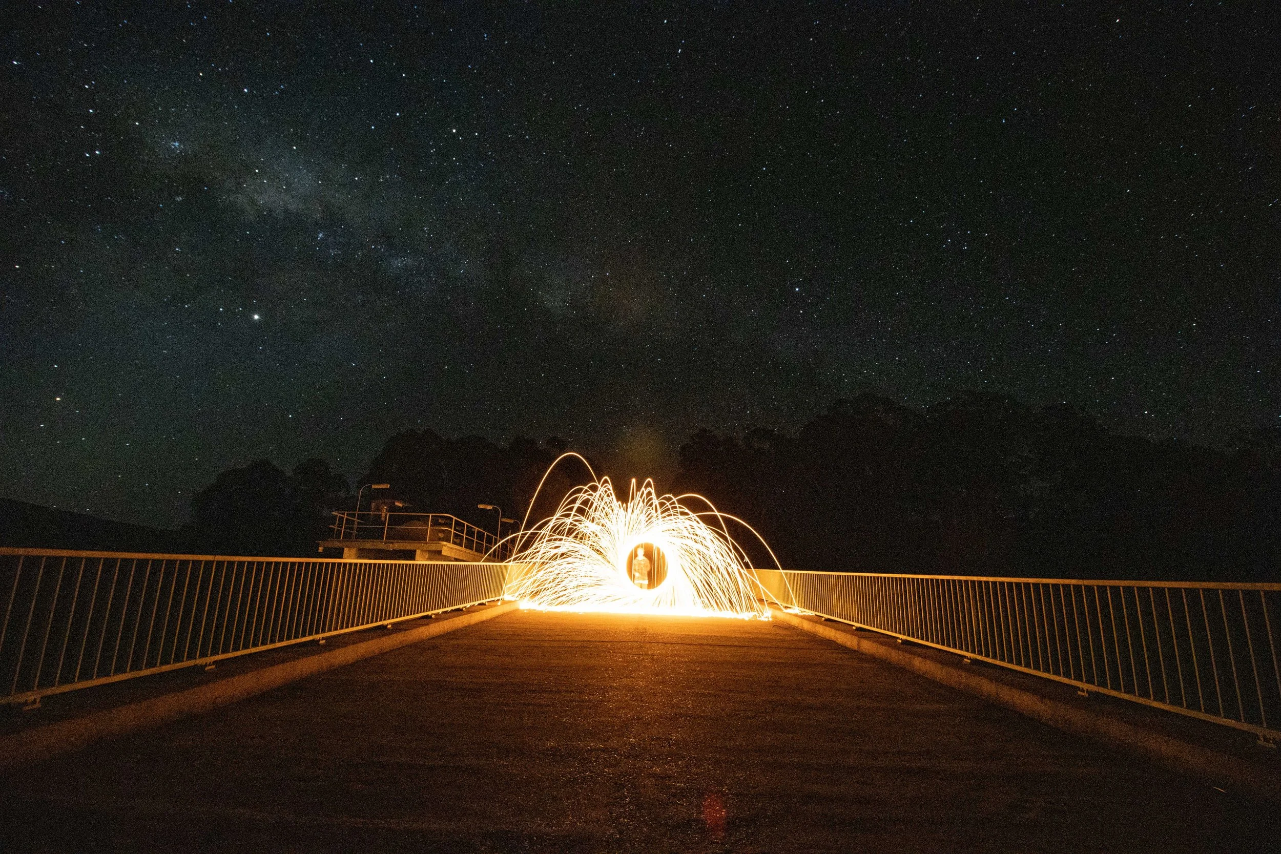 Person creating sparks with steel wool on a bridge at night, under a starry sky with the Milky Way visible.