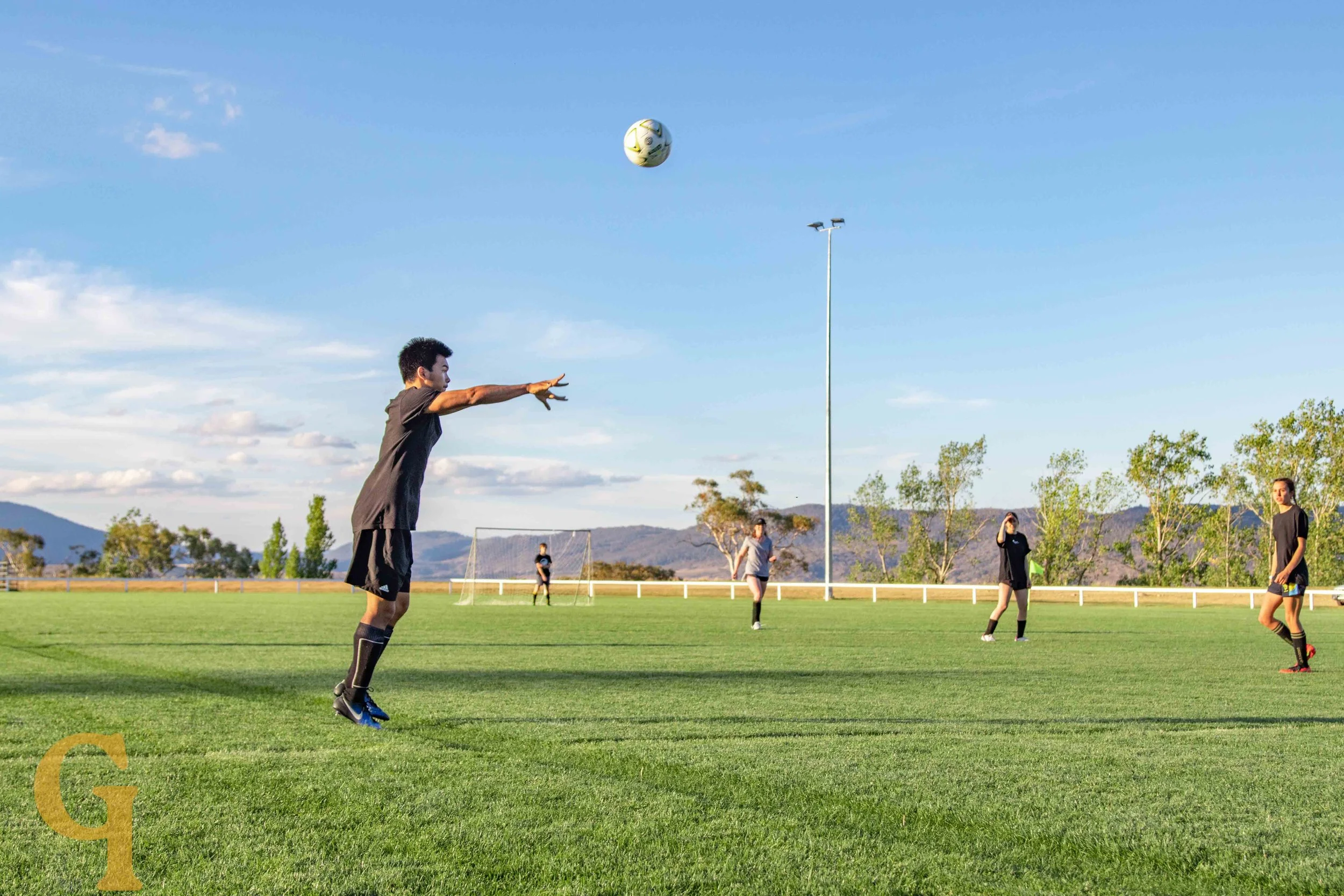 A group of women and a man playing soccer on a grassy field under a blue sky with some clouds, with mountains and trees in the background. One man is about to catch a soccer ball.
