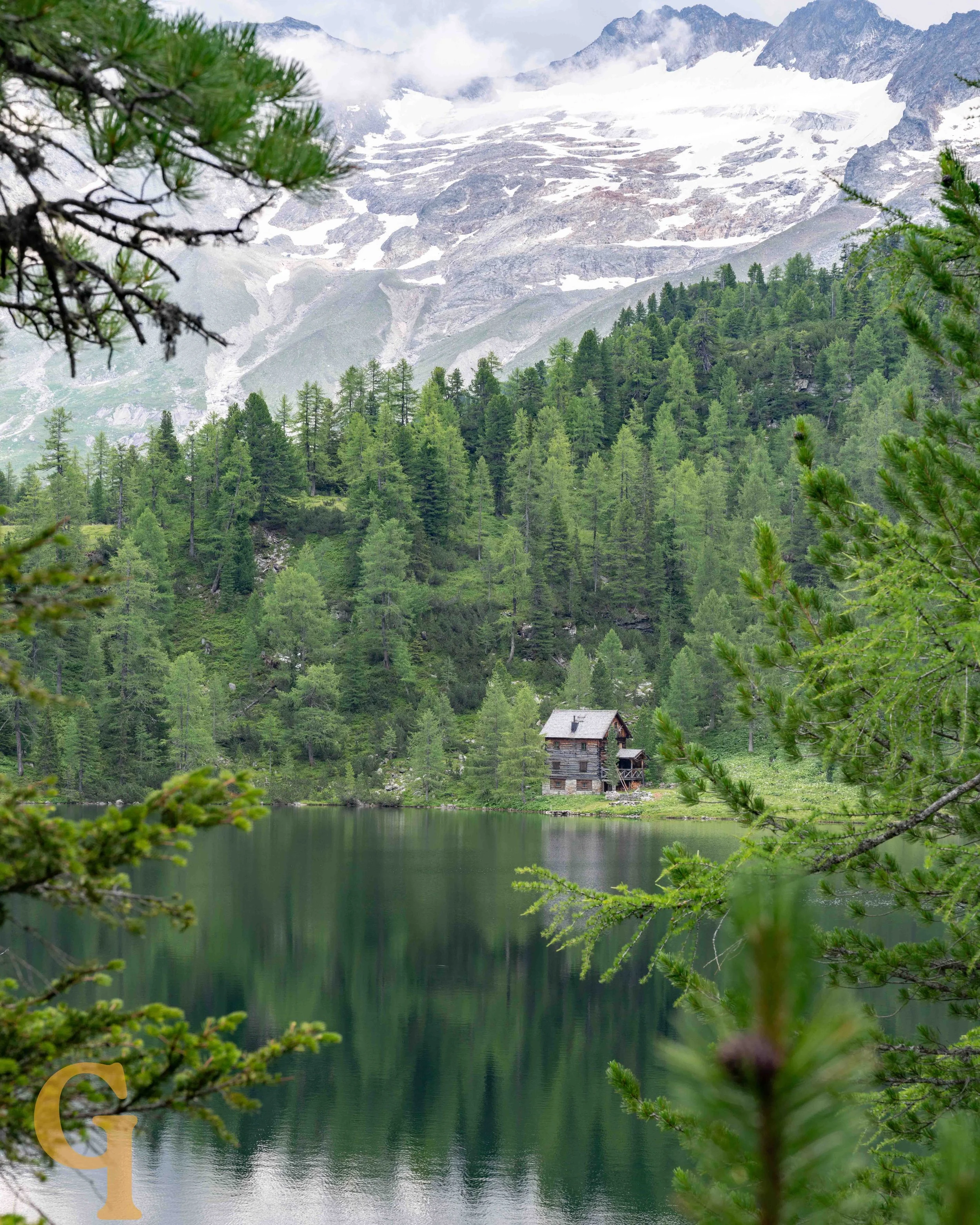A mountain lake surrounded by dense green pine trees with a rustic wooden cabin near the shore, snow-capped mountains in the background, and overcast sky.
