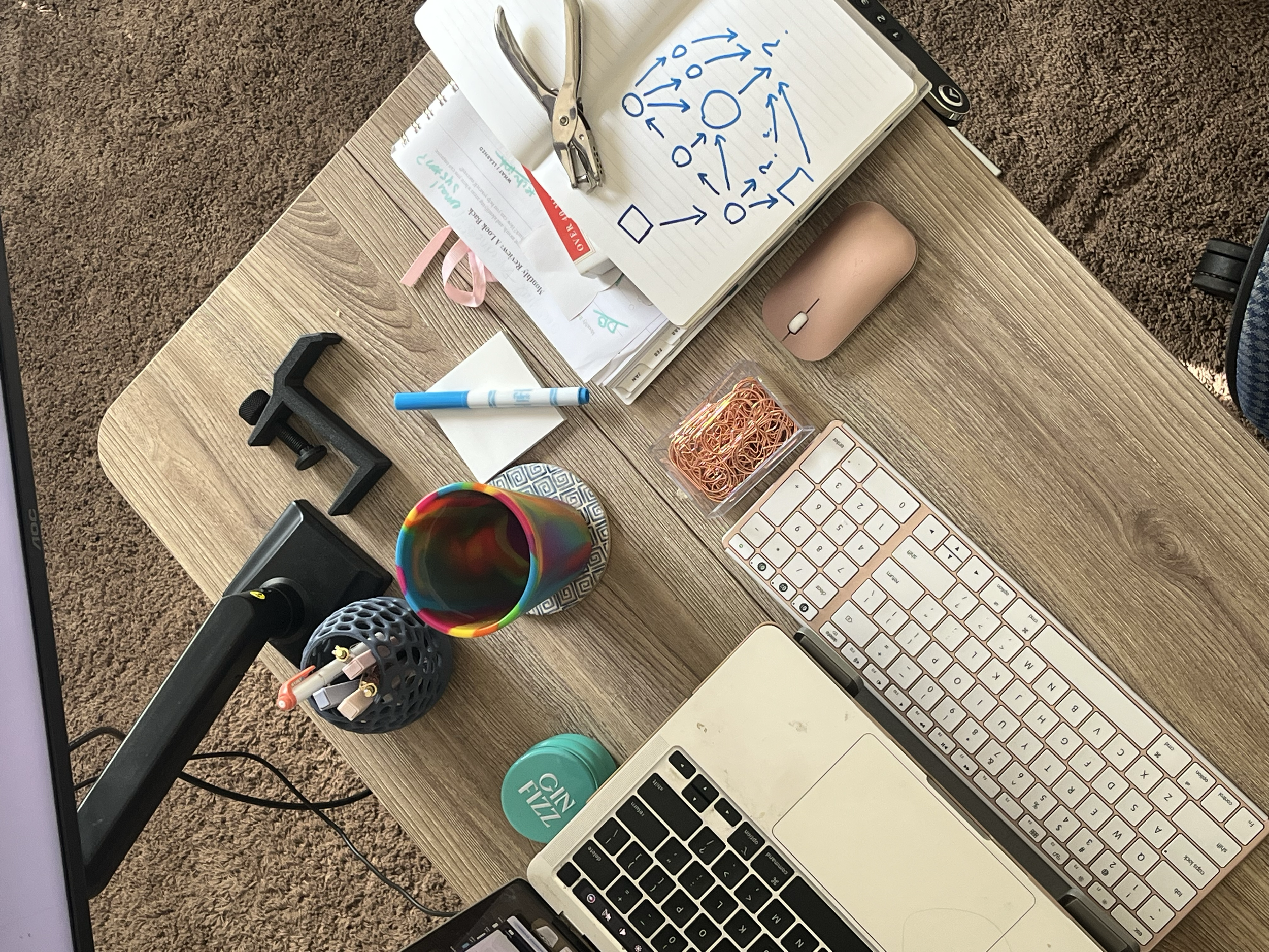 A wooden desk with a computer monitor, a keyboard, a mouse, a laptop, a rainbow-colored cup, a white notepad with a blue marker, several papers, a stapler, a container with pens and paper clips, and a coaster. The desk is on a brown carpeted floor.