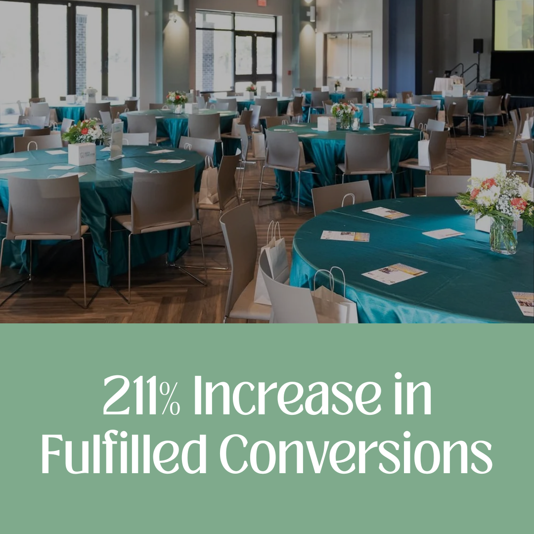 Event space with round tables covered in teal tablecloths, decorated with flower arrangements and place cards, in a well-lit room with large windows.
