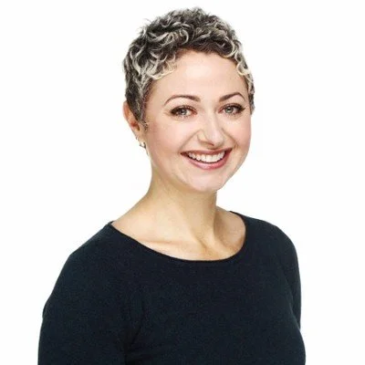 A woman with short, curly salt-and-pepper hair smiling, wearing a black top, against a white background.