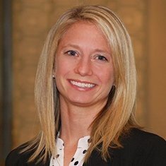 A woman with blonde hair smiling, wearing a dark blazer and a white top, against a neutral background.