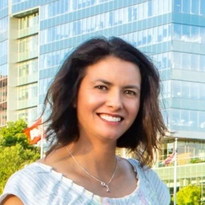 A woman with shoulder-length dark hair, smiling, standing outdoors in front of a modern glass building.