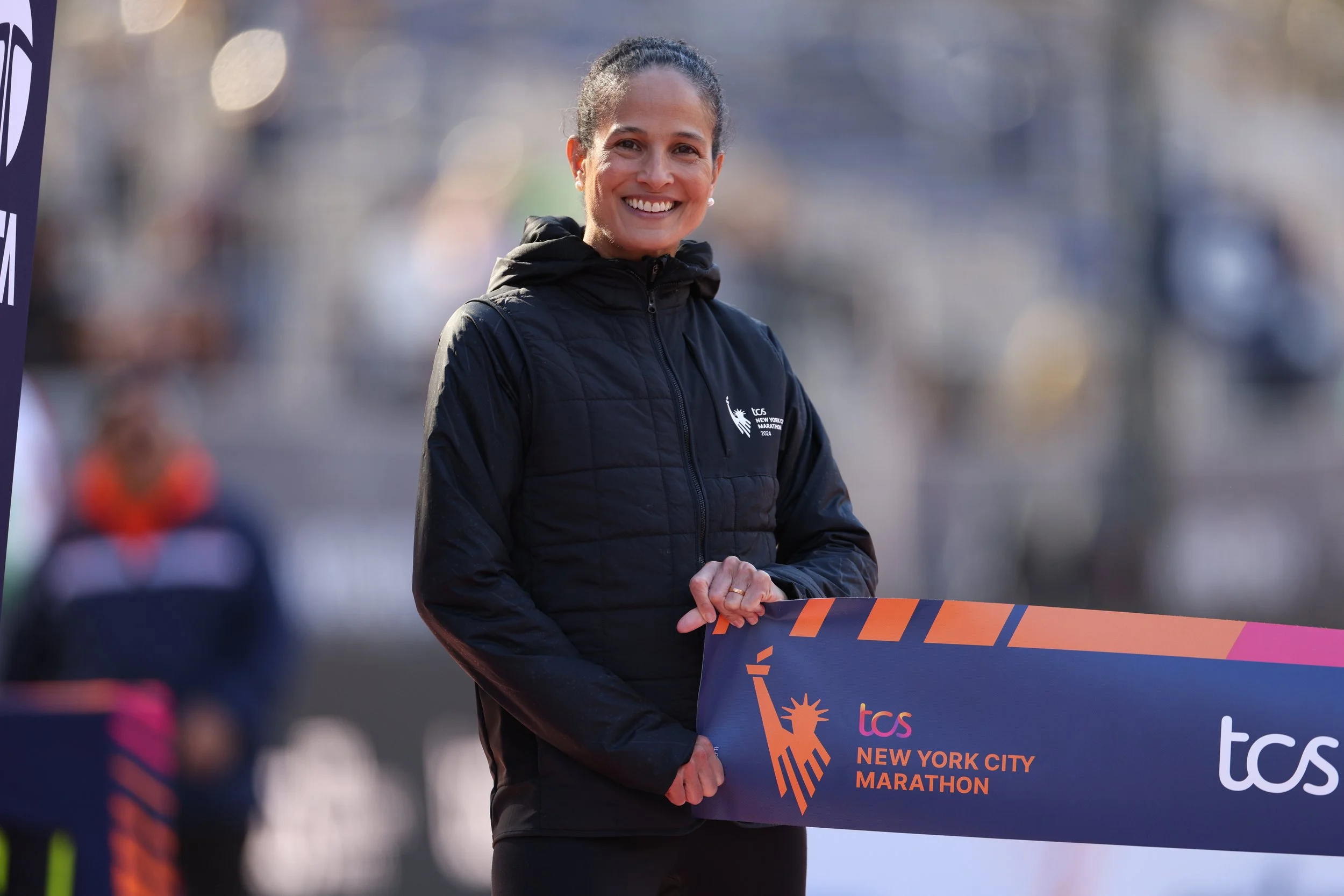 A smiling woman in athletic clothing holding a finish line banner at the TCS New York City Marathon.