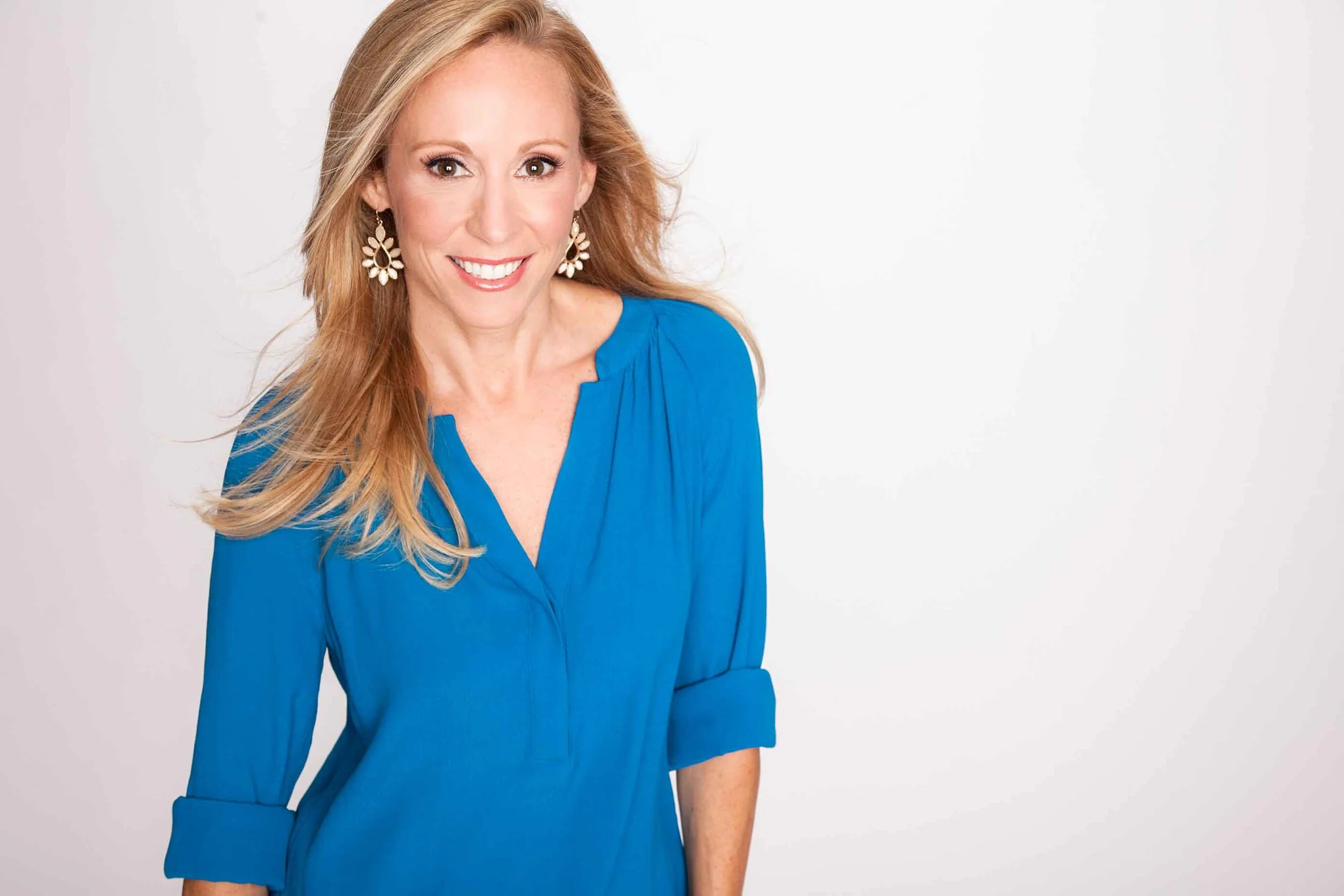A woman with long blonde hair wearing a blue dress and floral earrings, smiling against a plain white background.