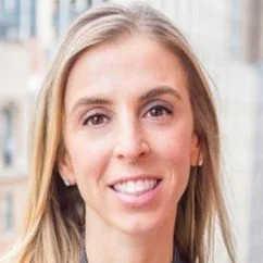 Close-up of a young woman with light brown hair smiling outdoors