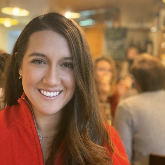 Smiling woman with brown hair wearing a red jacket in a crowded indoor setting, possibly a restaurant or cafe.