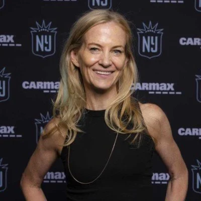 A smiling woman with blonde hair standing in front of a black backdrop with sports team logos.