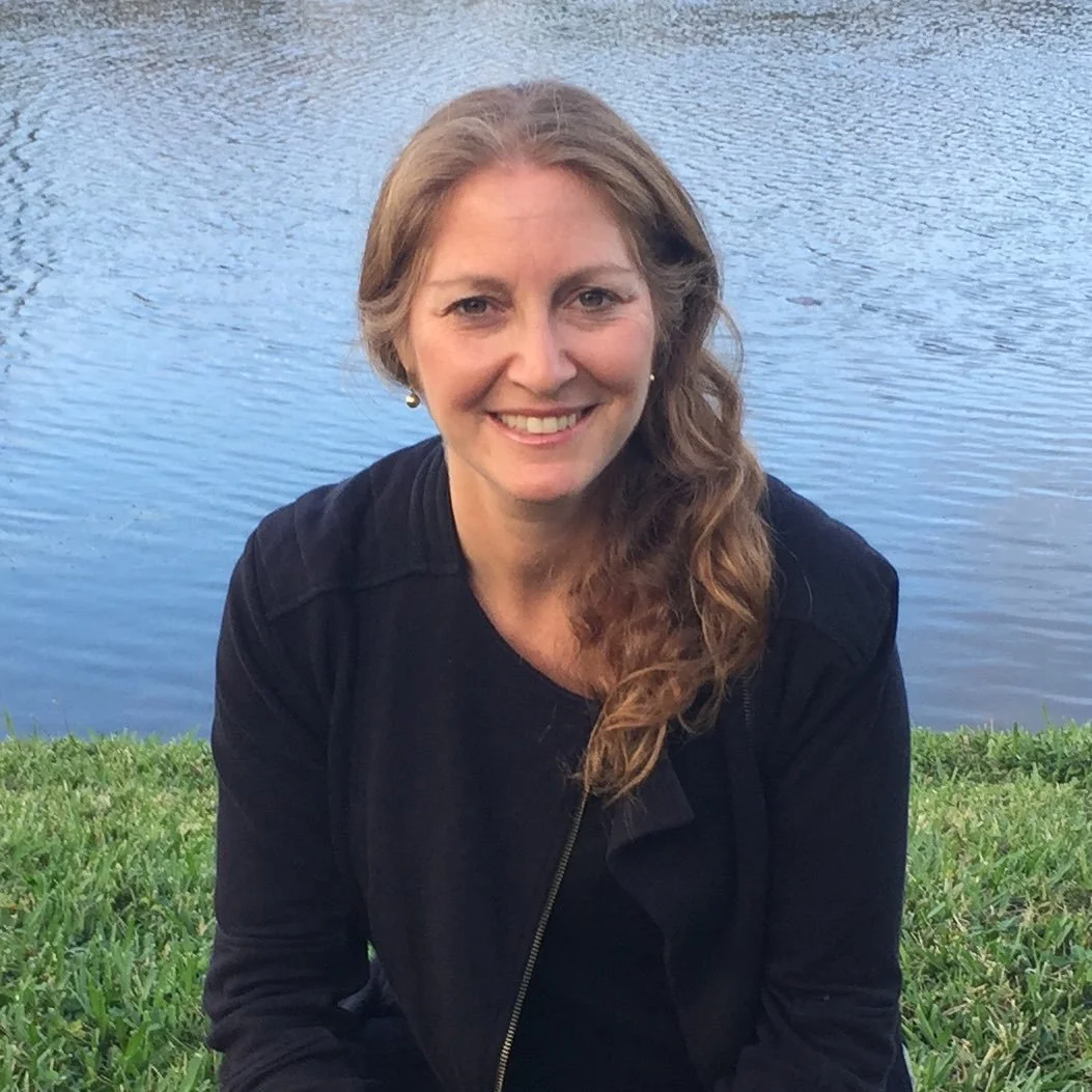 A woman with long, wavy, light brown hair smiling while sitting on grass near a body of water, wearing a black jacket and earrings.
