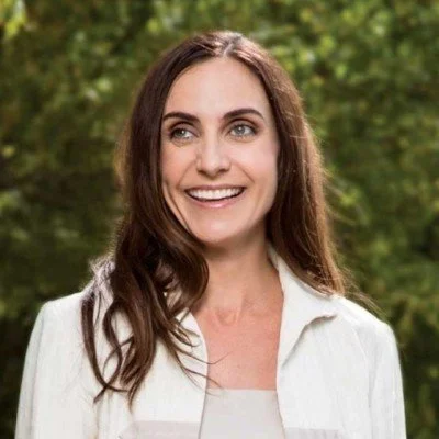 A woman with long brown hair smiling outdoors with green trees in the background.