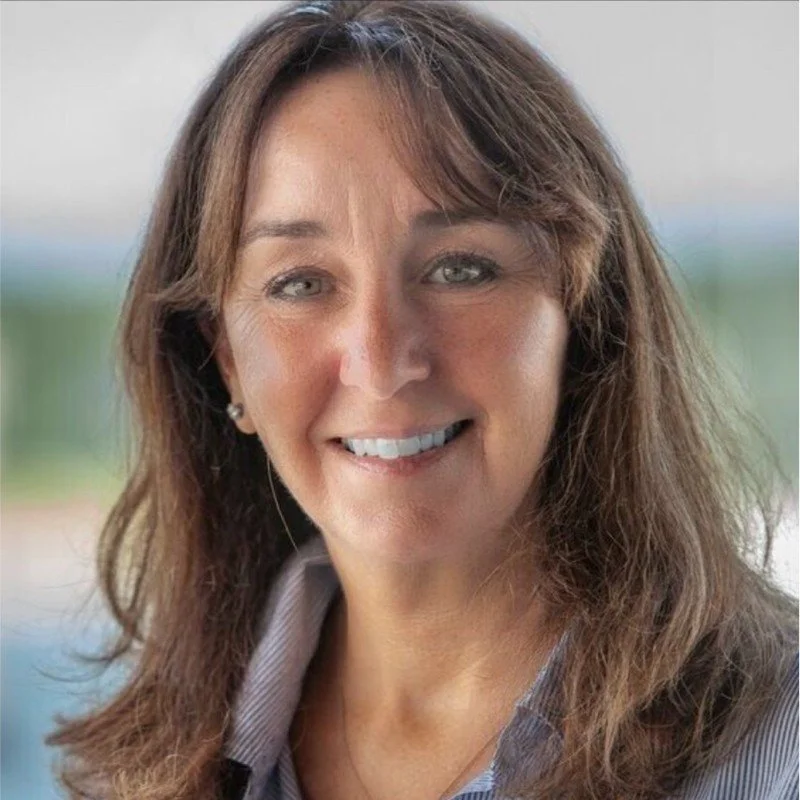 A woman smiling with shoulder-length brown hair, gray eyes, wearing pearl earrings and a collared shirt.