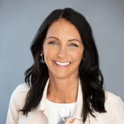 A woman with long black hair, wearing a white jacket and hoop earrings, smiling against a light gray background.