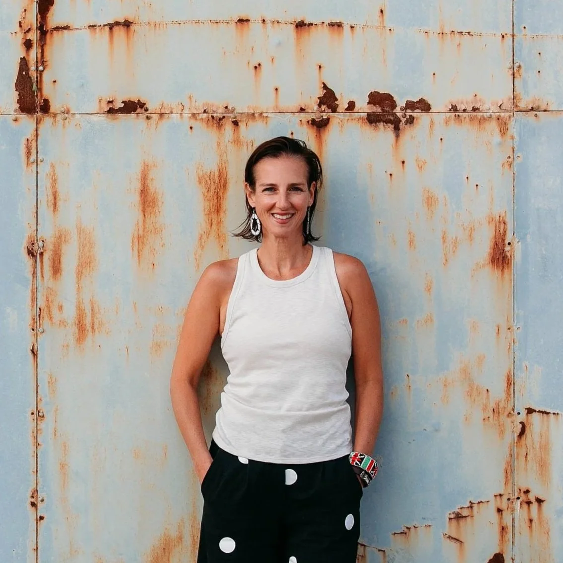 A woman with short dark hair smiling and standing in front of a weathered, rusty metal wall. She is wearing a sleeveless white top, black pants with large white polka dots, colorful earrings, and a beaded bracelet.