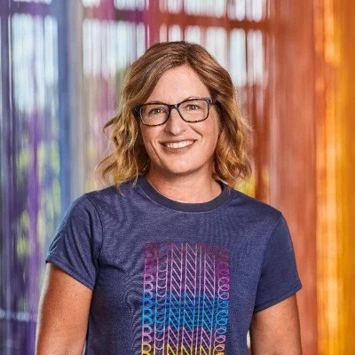 A smiling woman with curly blond hair and glasses, wearing a blue t-shirt with colorful geometric text, standing in front of vibrant, multicolored curtains.