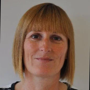 Close-up portrait of a woman with straight, shoulder-length red hair and bangs, smiling slightly against a plain background.