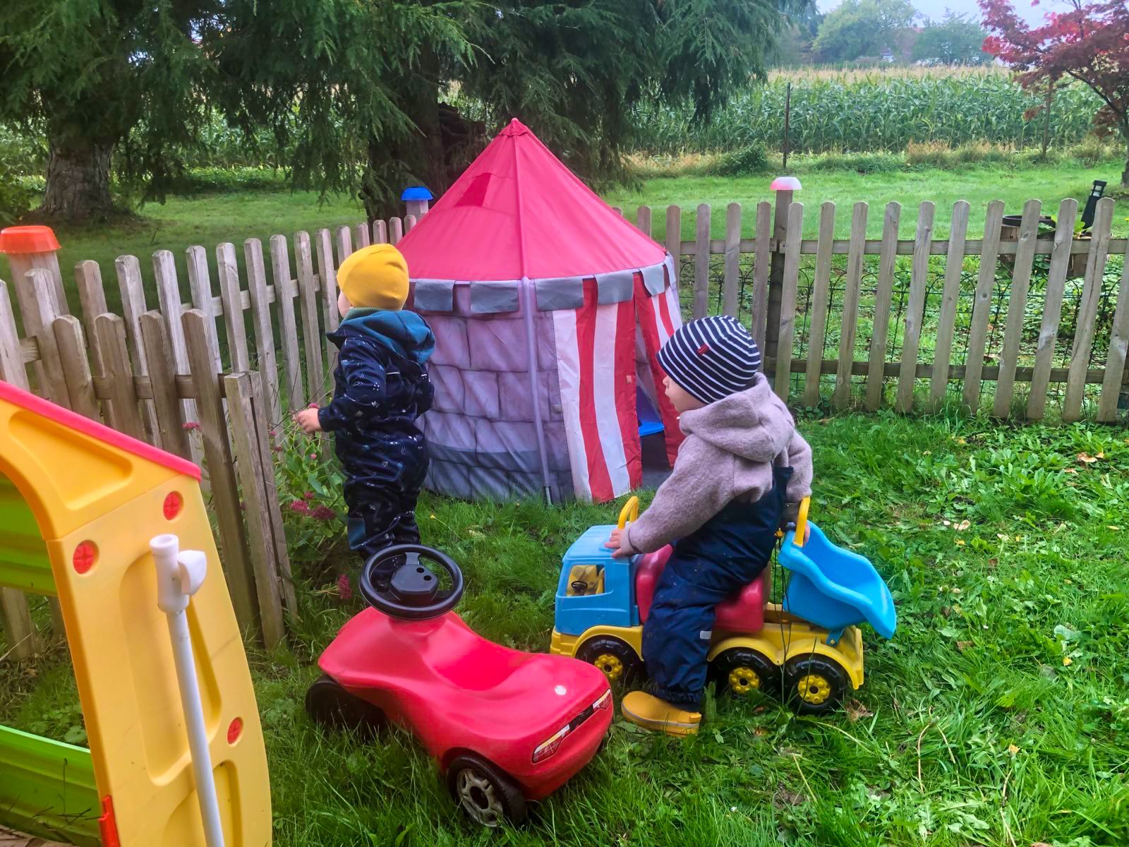 Zwei Kinder spielen im Garten, eines vor einem roten Spielzeugauto und das andere bei einer bunten Zirkus- oder Trommelzelte.