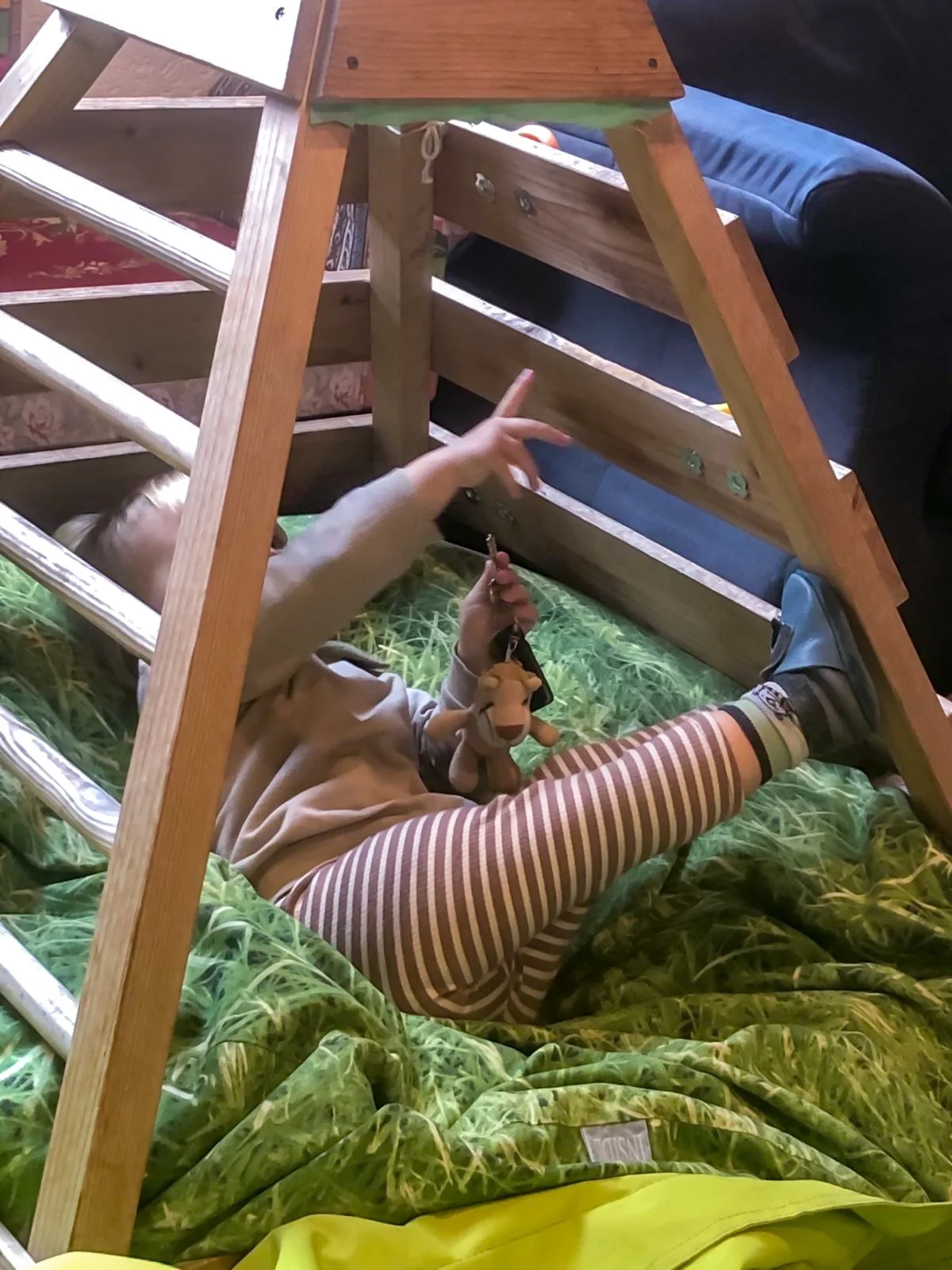 Ein Kind liegt unter einem Klettergerüst auf einer grünen Decke, hält einen Schlüsselbund mit einem Plüschtier und zeigt nach oben.
