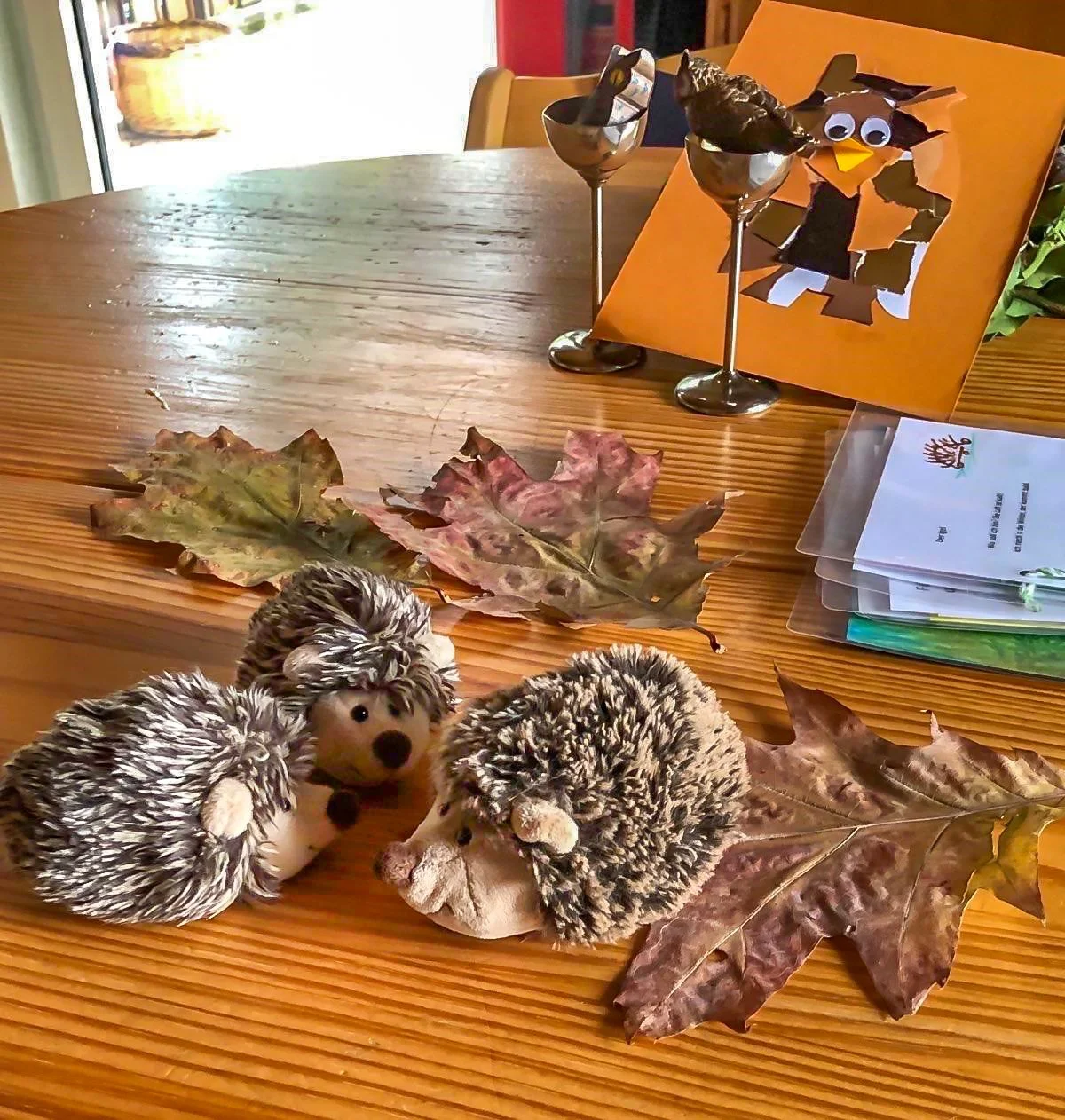 Herbstdekoration auf einem Holztisch mit getrockneten Blättern und drei Plüsch-Igeln. Im Hintergrund ein orangefarbenes Kunstwerk mit einer Eule aus Papierschnipseln.