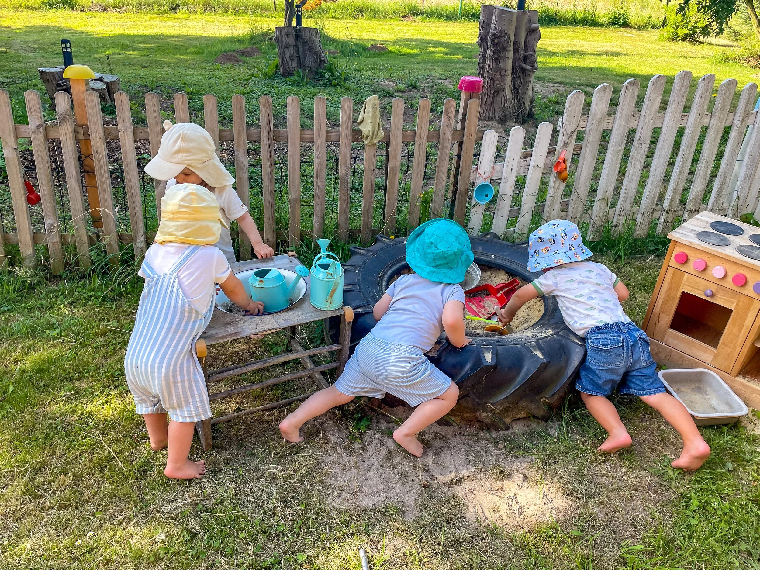Vier Kinder spielen draußen im Sandkasten, umgeben von einer Holzumzäunung, mit Spielzeugküche und Gartengeräten, bei sonnigem Wetter im Garten.