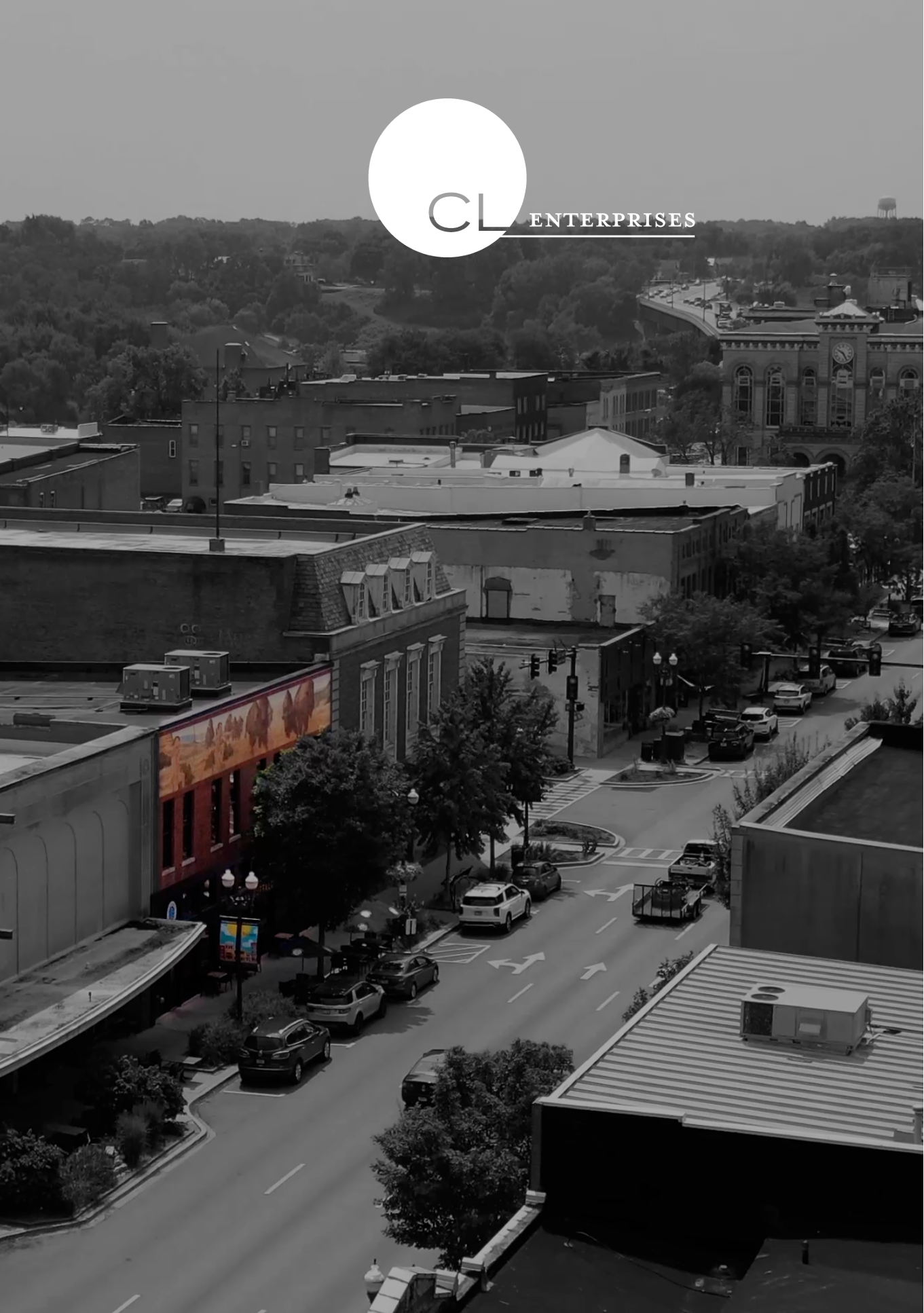 A black and white photo of a small downtown area with parked cars, trees, and buildings. In the background, there are hills with a water tower and a bridge with traffic.