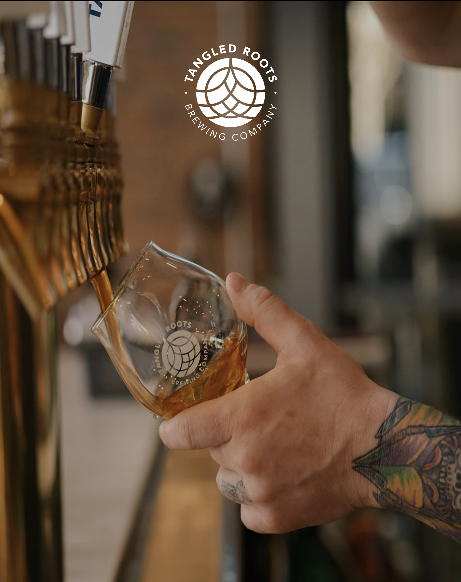 Person pouring beer from tap into a glass at a brewery, with a tattooed arm and colorful sleeve visible.